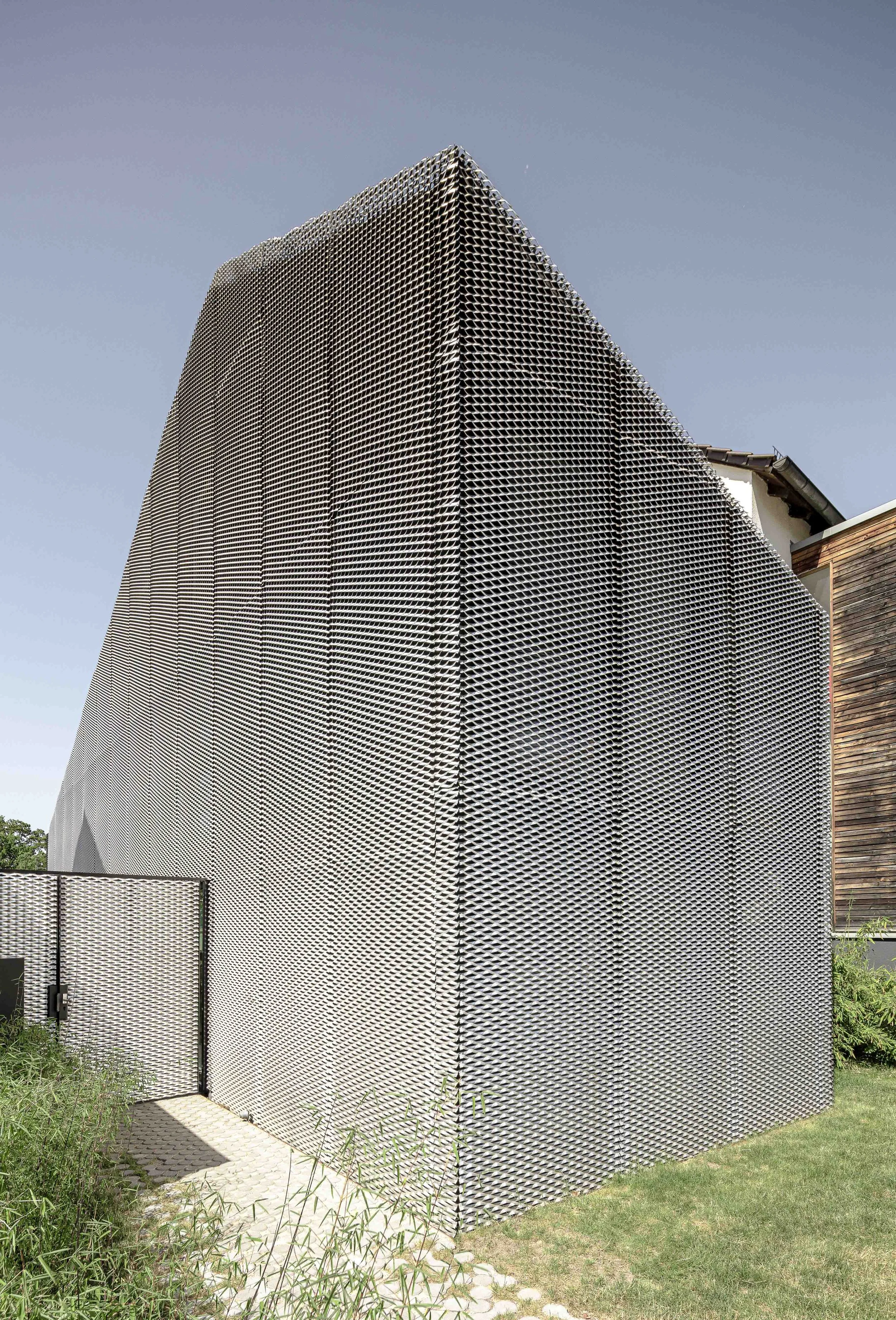 Ein modernes Gebäude mit einer perforierten metallisierten Fassade, das an eine Klippe oder eine Struktur mit einer texturierten Oberfläche erinnert. Das Gebäude ist von grünem Gras und Büschen umgeben, mit einem klaren blauen Himmel im Hintergrund.
