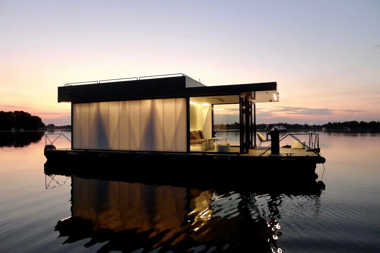 loungeboat - Modernes Hausboot auf ruhigem Wasser bei Sonnenuntergang, mit sichtbarem Innenraum.