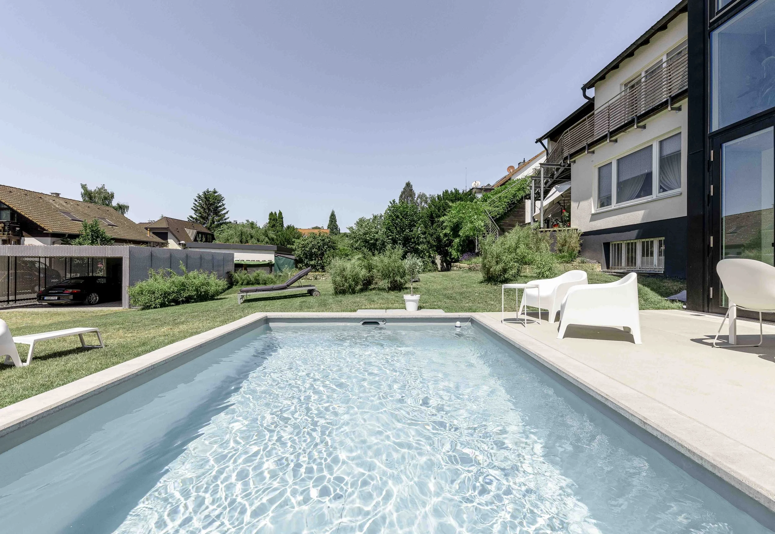 Garten mit Swimmingpool, Sonnenliegen, runden Tisch, Stühle, Bäume und Häuser im Hintergrund, klarer blauer Himmel.