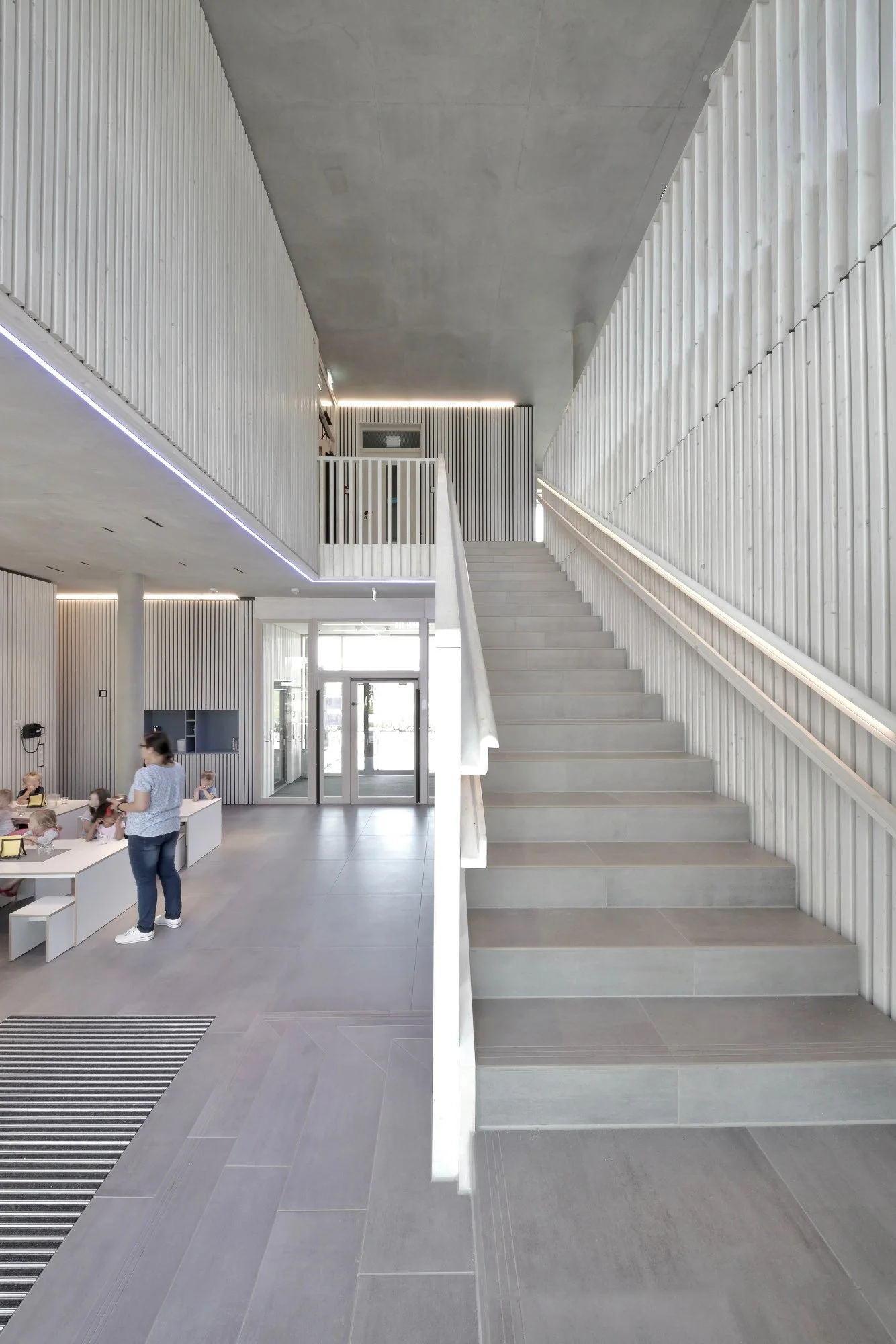 kinderhaus BUGA - Innenraum eines modernen Gebäudes mit grauen Fliesen, einer weiß gestrichenen Treppe und vertikalen Holzverkleidungen an den Wänden. Im Hintergrund sind Kinder und eine erwachsene Person in einem hellen Raum sichtbar.