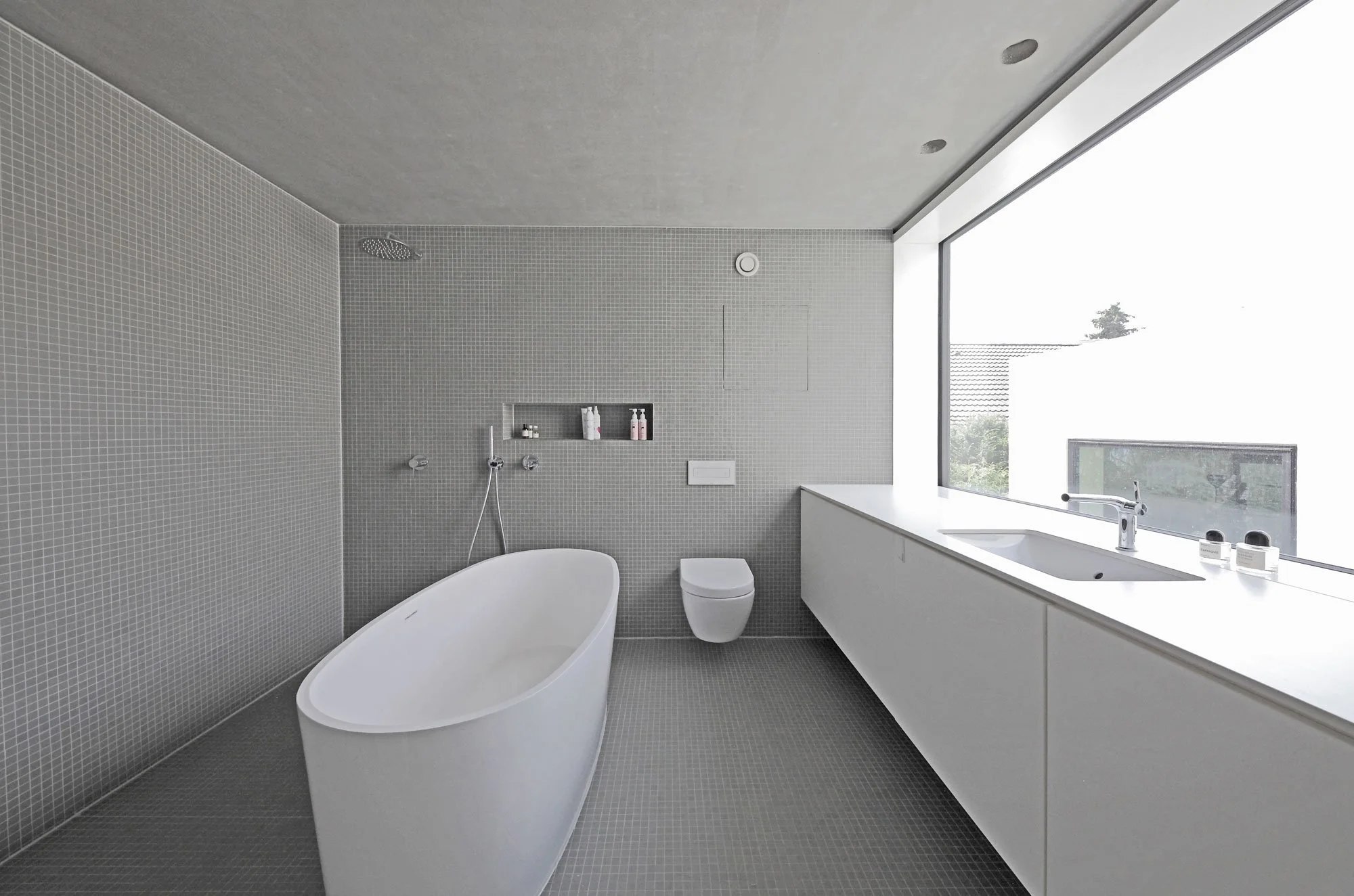 haus SG - moderner, minimalistischer Badezimmer mit Badewanne, WC, Waschbecken, großem Fenster und grauen Fliesen.