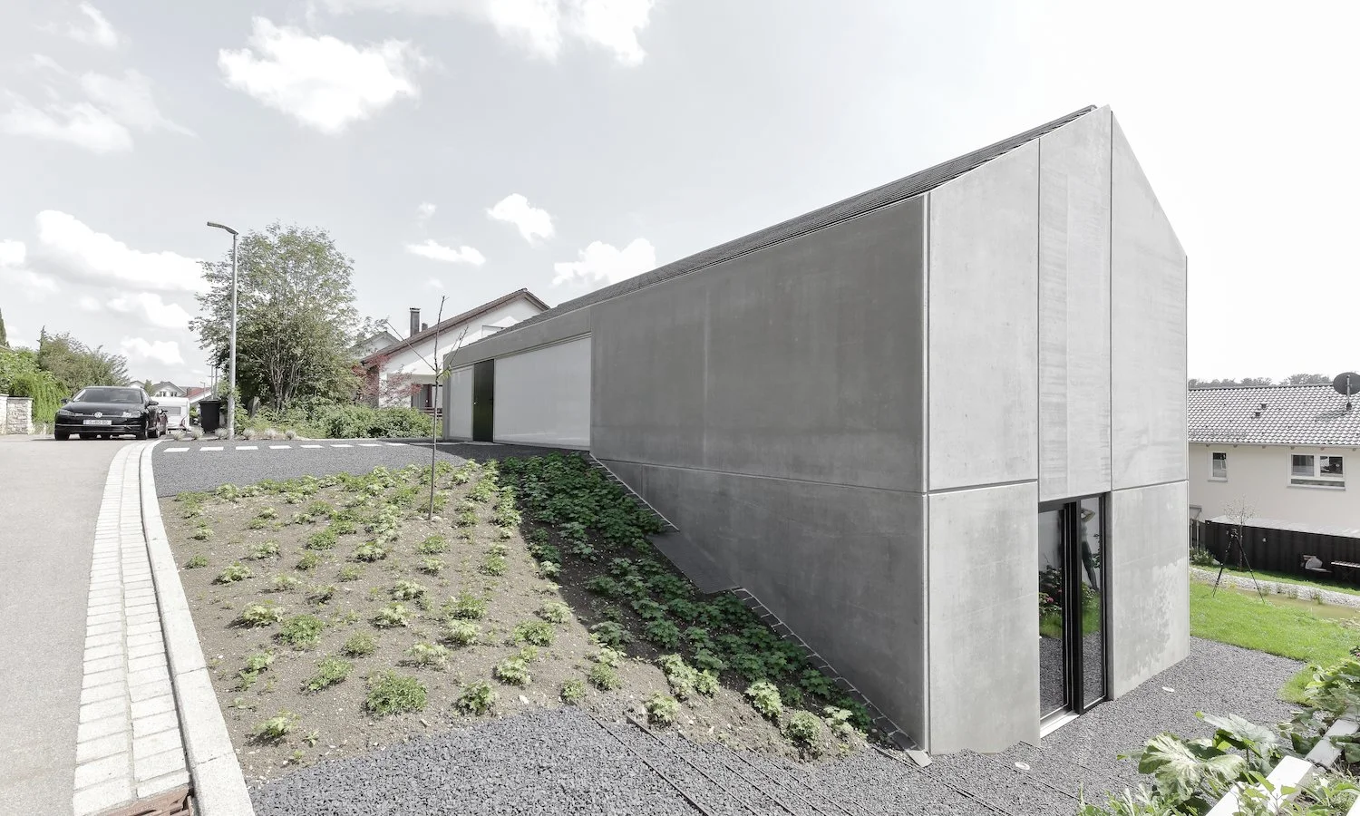 haus WÄ - Moderne, zweistöckige graue Betonhaus mit schrägem Dach, großes Fenster und Gartenterrasse in einem Vorort, umgeben von kleineren Häusern und Bäumen.
