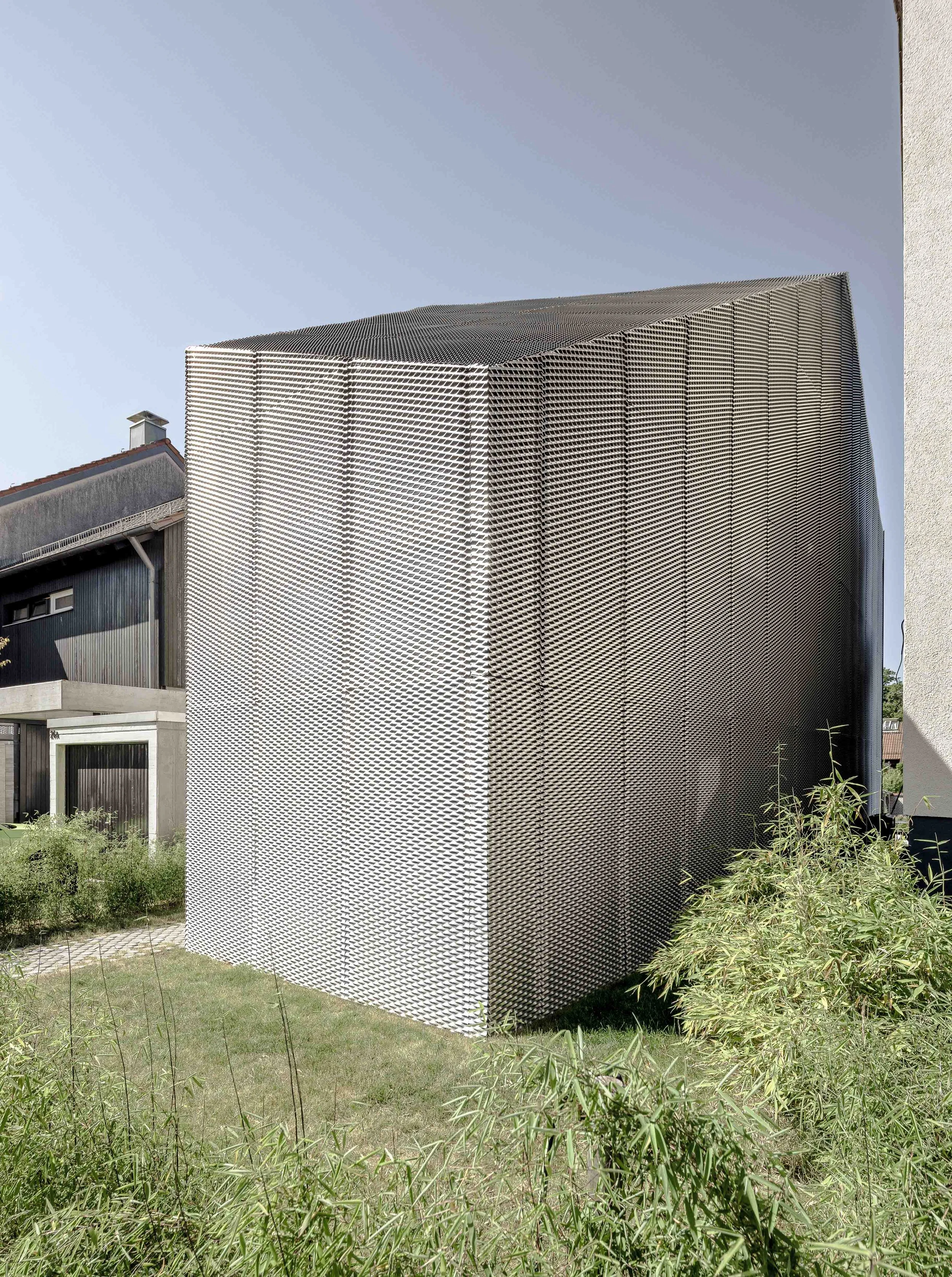Moderne Gebäude mit metallischer Außenfassade aus arbeitsspezifischen, horizontalen Schlitzen, umgeben von Pflanzen und einem gekachelten Weg. Der Himmel ist klar und blau.
