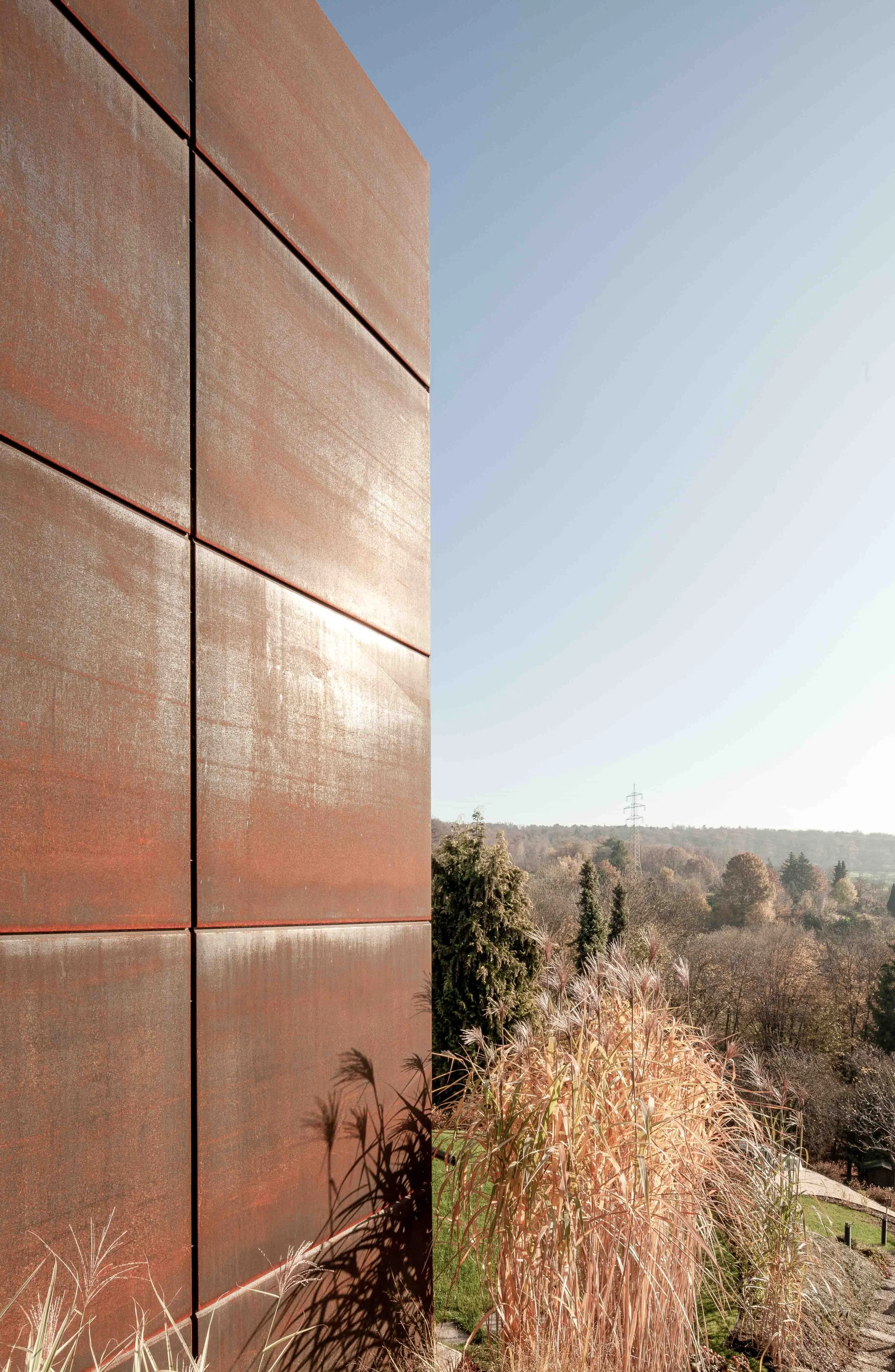 haus GRO - Ein modernes Gebäude mit roten Metallplatten an der Außenwand, umgeben von Büschen und Bäumen, mit einer Landschaft im Hintergrund und einem blauen Himmel.