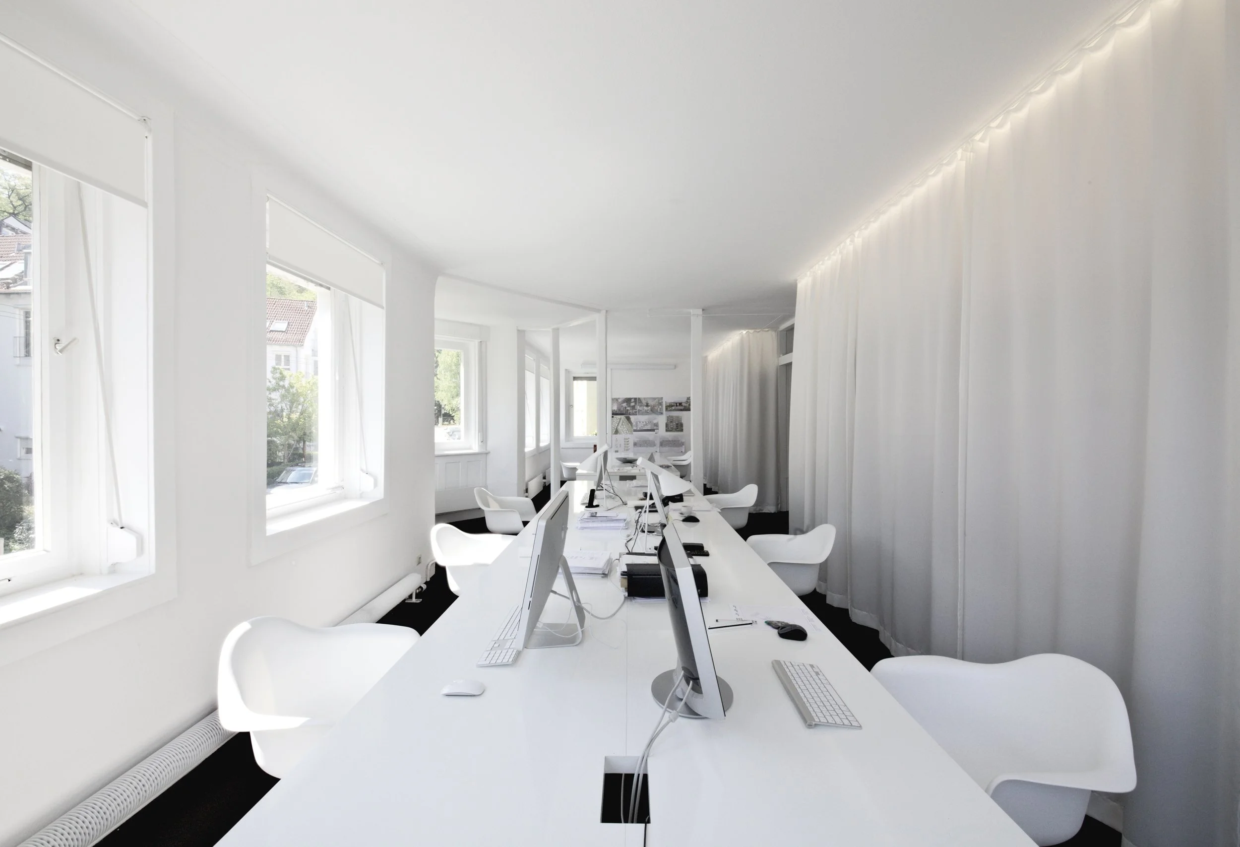 Ein heller, moderner Büroraum mit mehreren weißen Tischen und Computern, großen Fenstern mit weißen Jalousien und weißen Stühlen, gut beleuchtet durch Tageslicht.