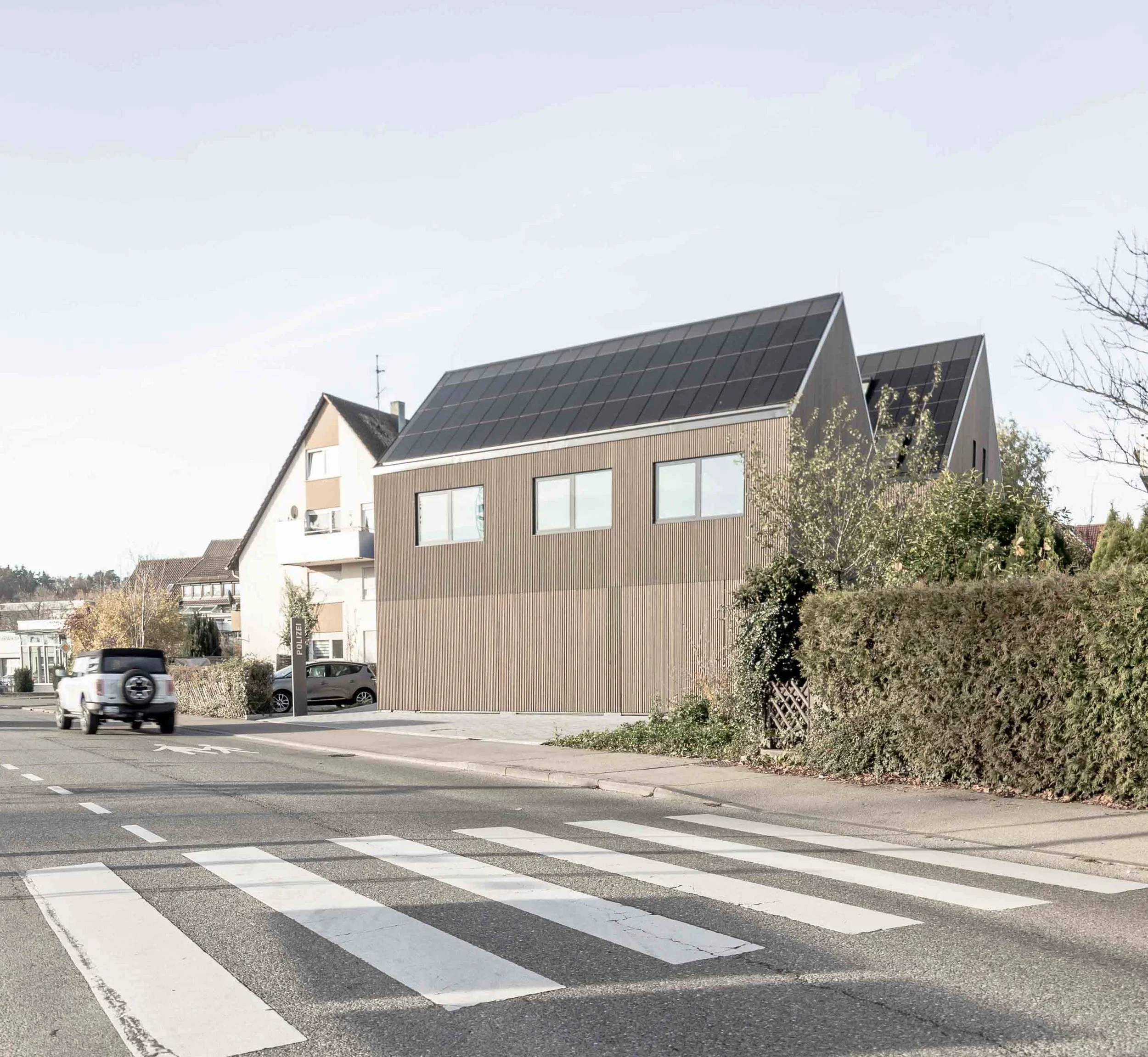 polizeiposten LE - Straßenkreuzung mit Zebrastreifen, parkende Autos, moderne Gebäude im Hintergrund, Baum und Busch im Vordergrund, blauer Himmel