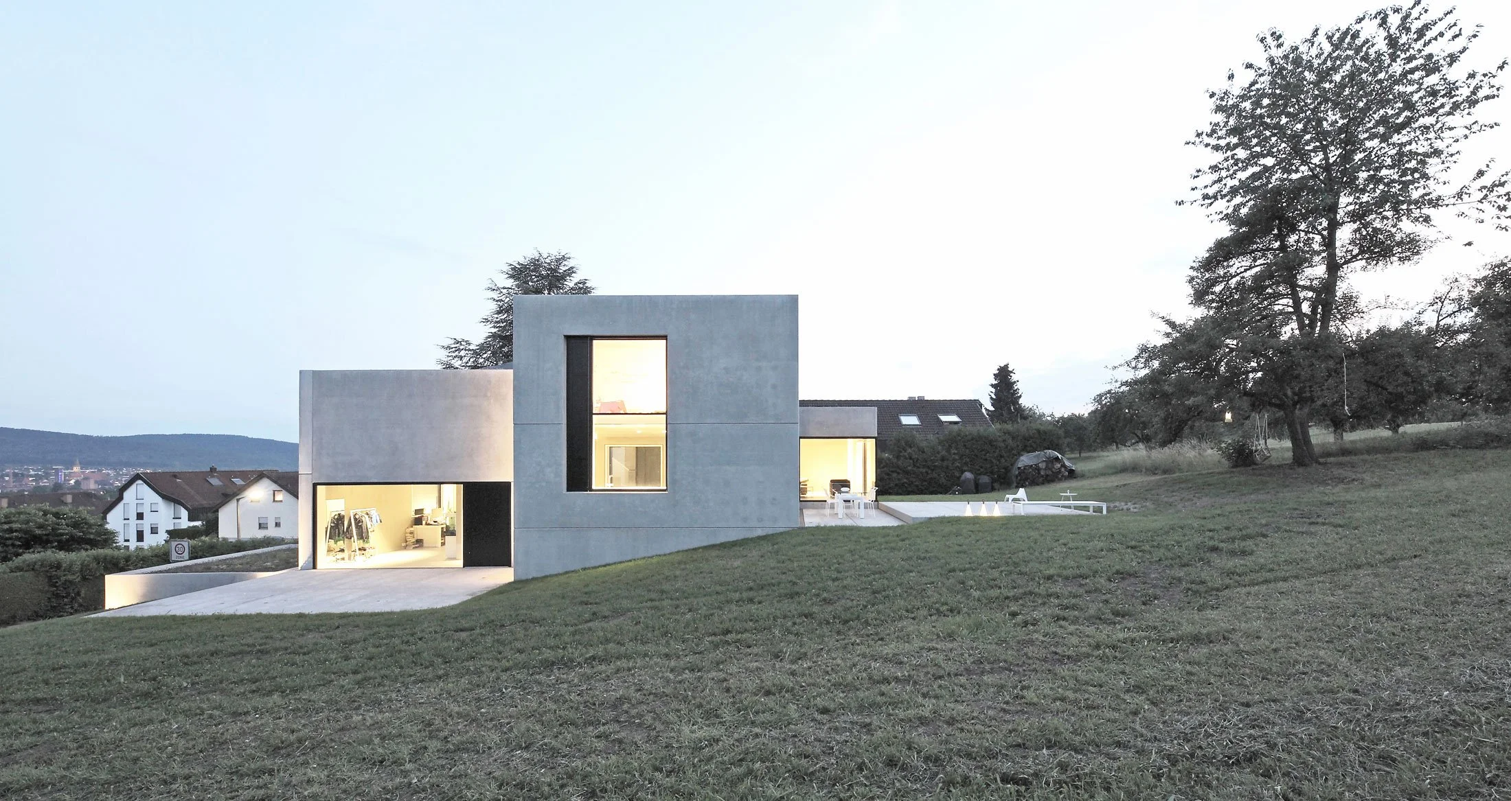 haus SG - Moderne, kubische Hausarchitektur mit großen Fenstern, auf einem Hügel bei Sonnenuntergang, umgeben von Bäumen und niedrigem Gras, in einer ländlichen Umgebung.