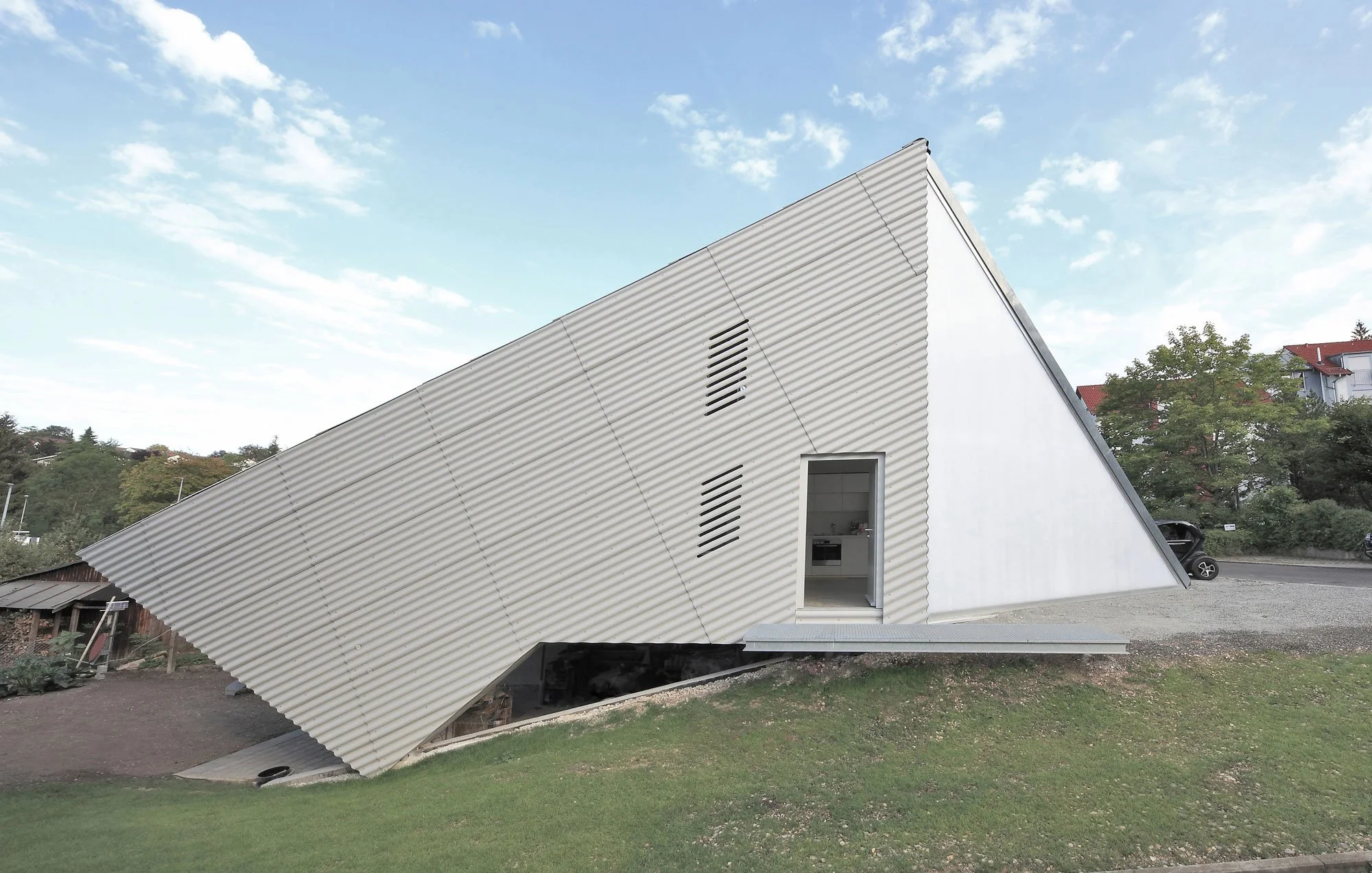 haus SO - Ein modernes, schräg gebautes Haus mit einer Kombination aus weißen Wänden und metallischer Fassade. Es hat eine offene Tür im Erdgeschoss und große, schmale Belüftungsschlitze hoch am Haus. 