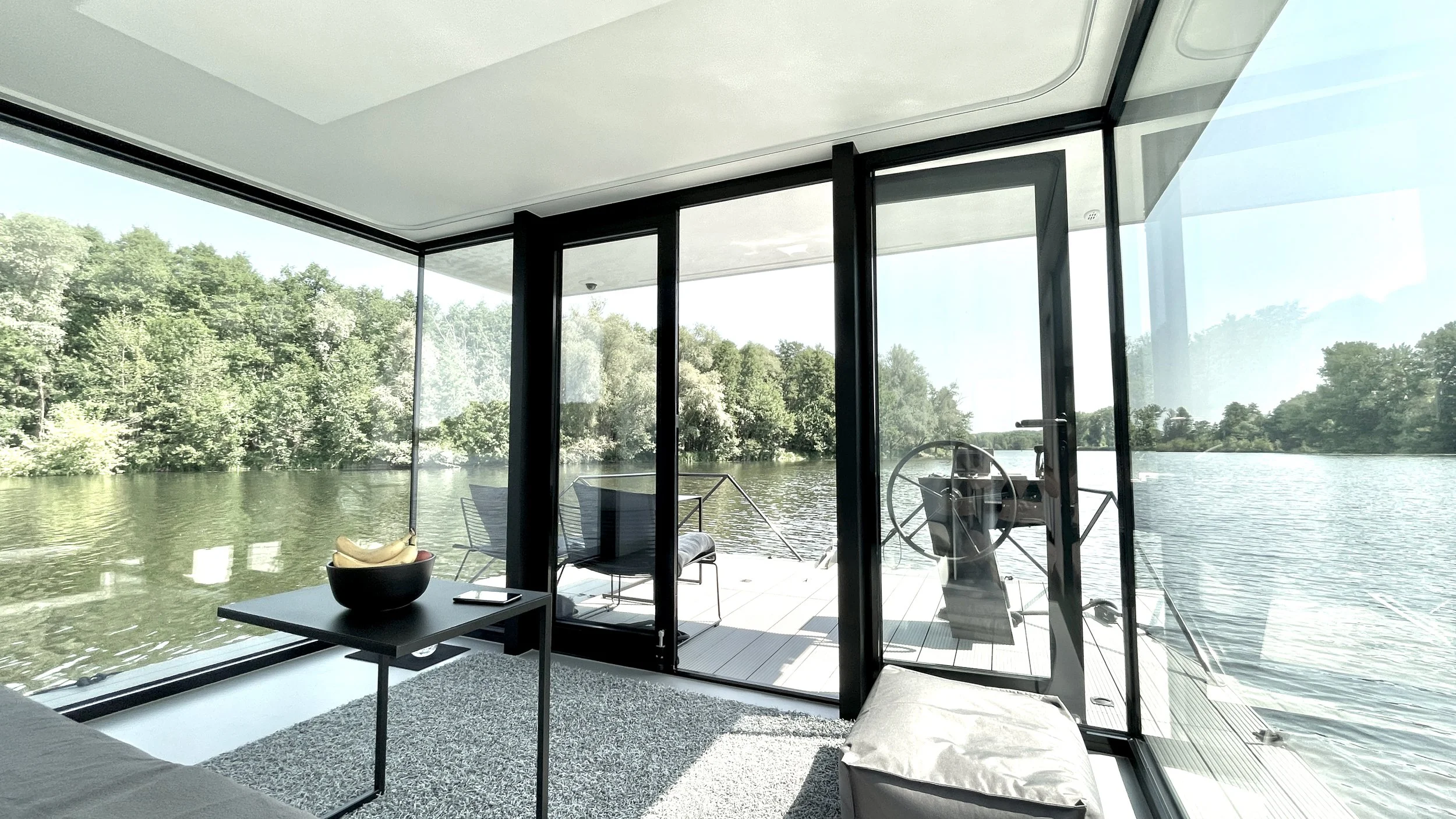 Moderne Wohnung am Wasser mit großen Fenstern und Blick auf einen Fluss, eingerichtet mit Stühlen, Tischen und einem Steuerrad auf einem Balkon.