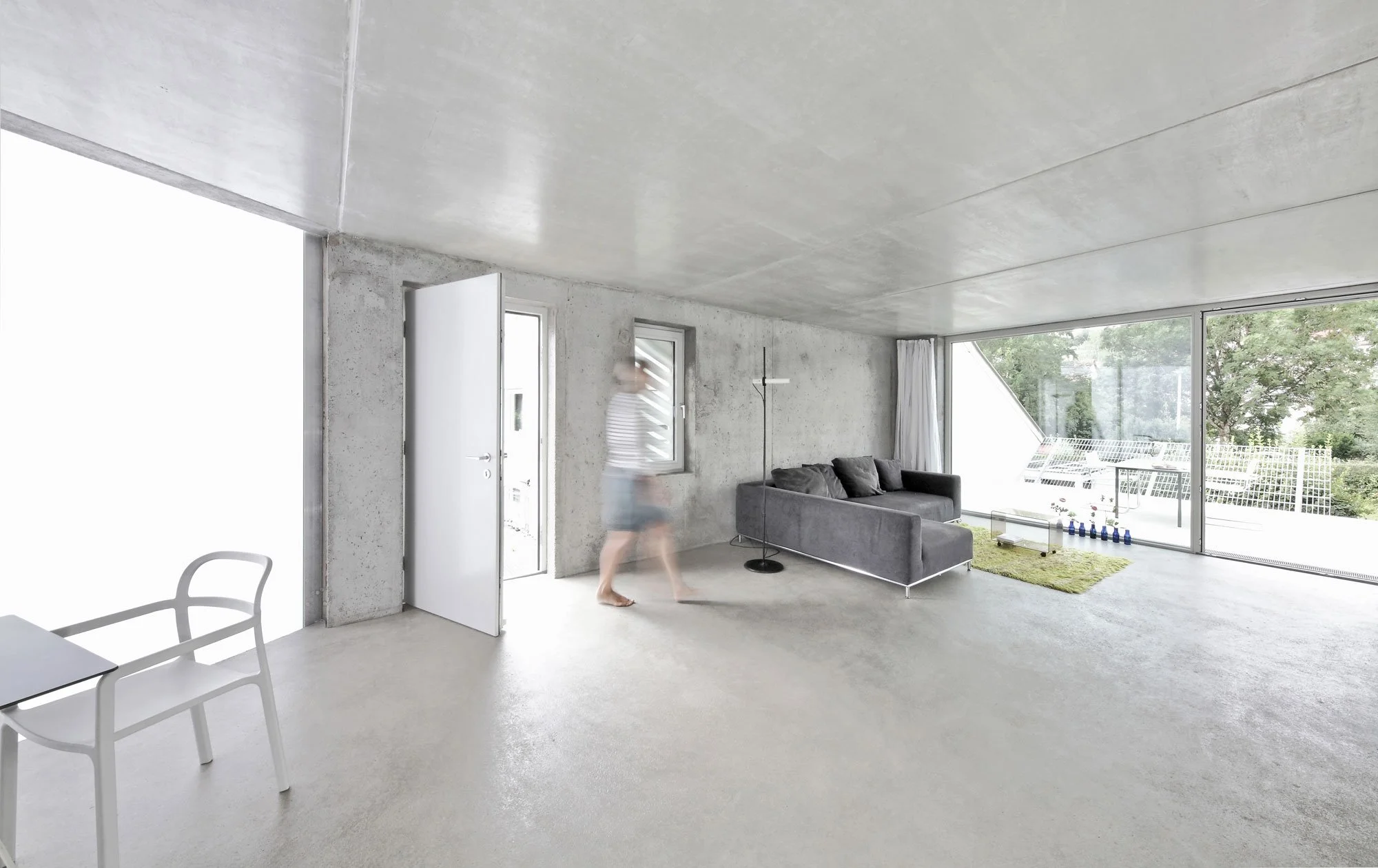 haus SO - Innenaufnahme eines modernen, minimalistischen Wohnzimmers mit großen Fenstern, einem grauen Sofa, einem weißen Couchtisch, einer Stehlampe und einem grünen Teppich, mit einer Person, die barfuß im Raum geht.