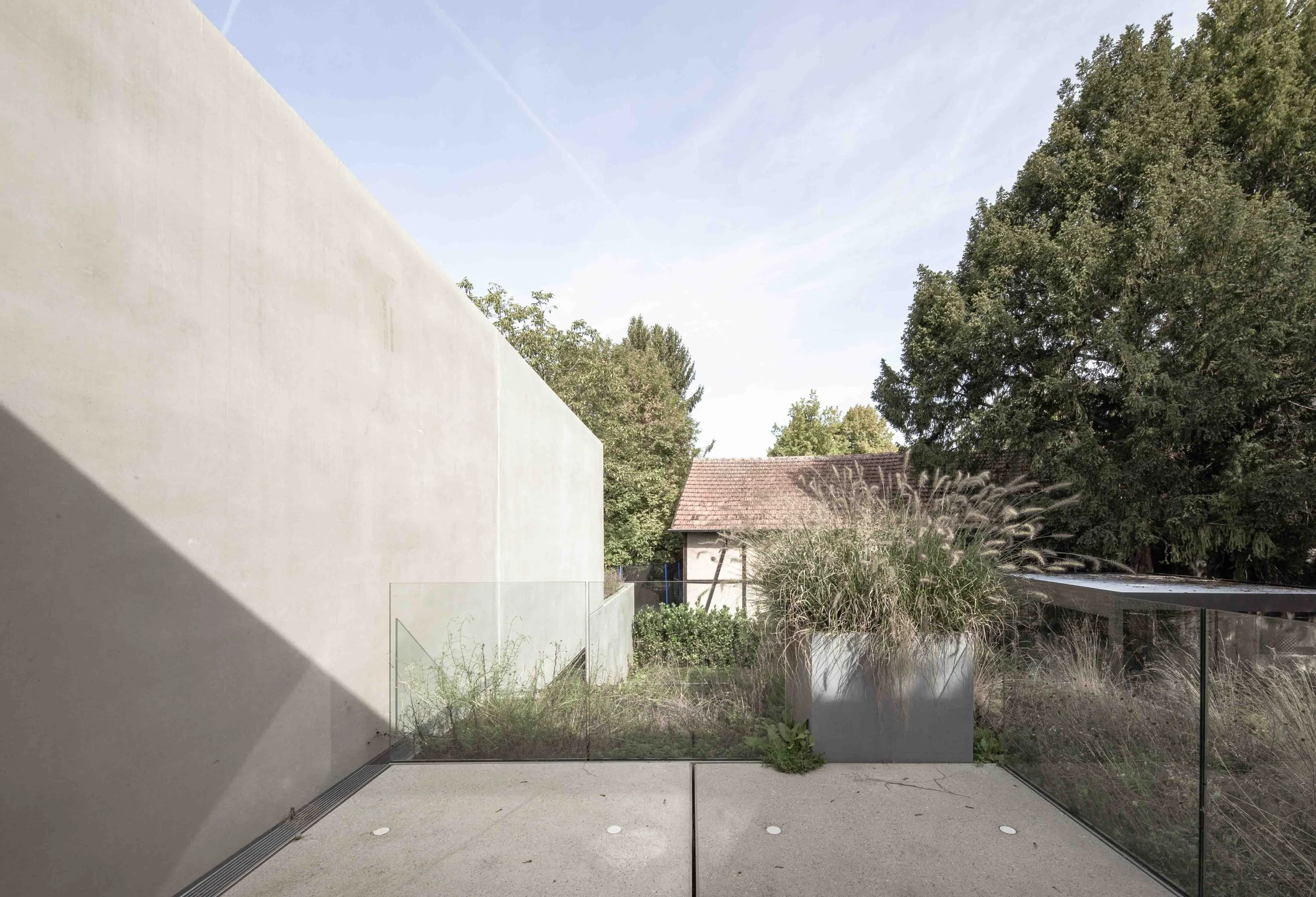 haus vO - Blick durch eine moderne, große Fensterwand auf einen Garten mit Bäumen, Gras und einem roten Gartenschuppen in einer Nachbarschaft.