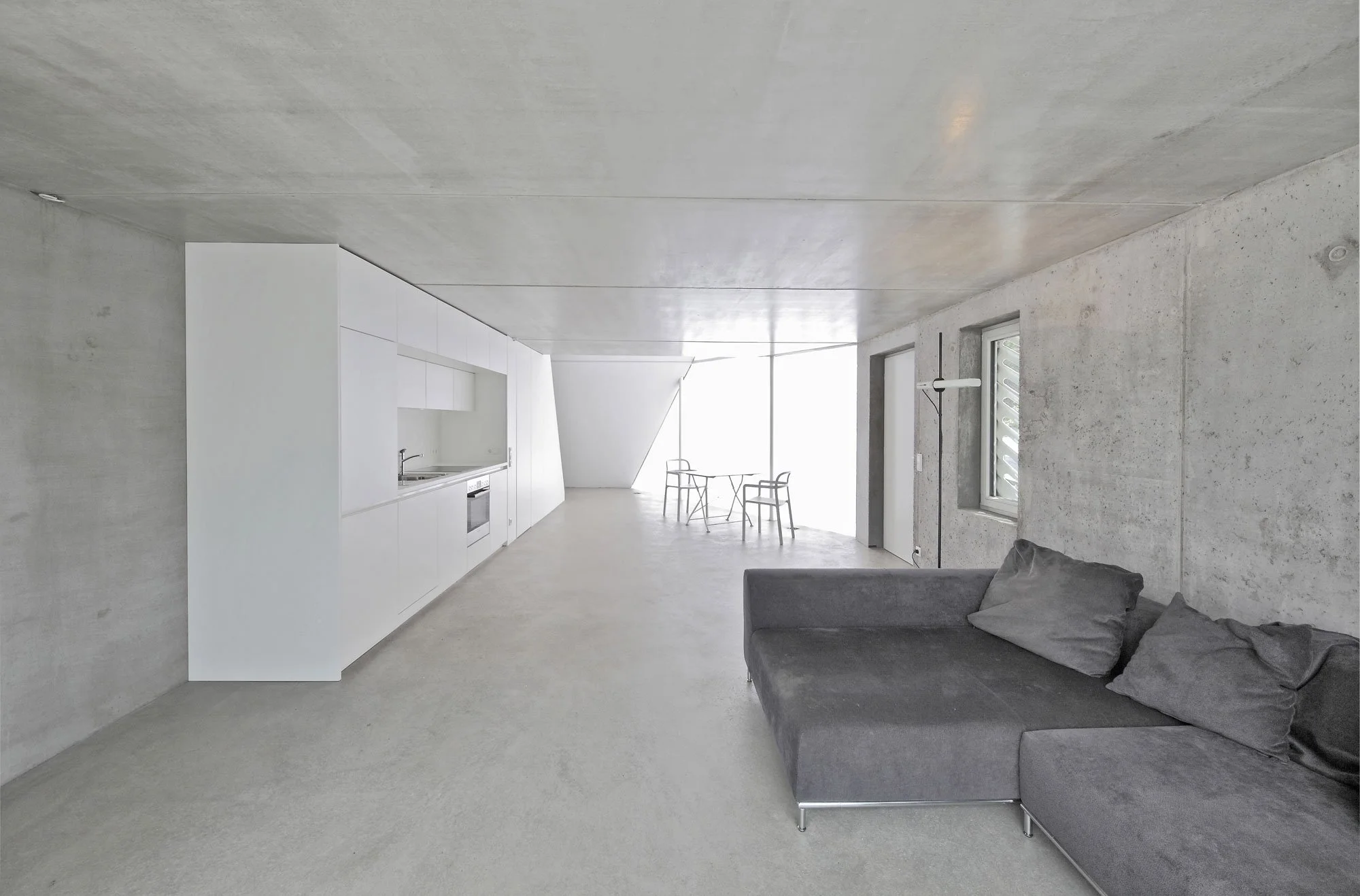 haus SO - Ein modernes, helles Wohnzimmer mit einer schwarzen Couch, grauen Wänden, einem weißen Küchenbereich und großen Fenstern, die natürliches Licht hereinlassen.