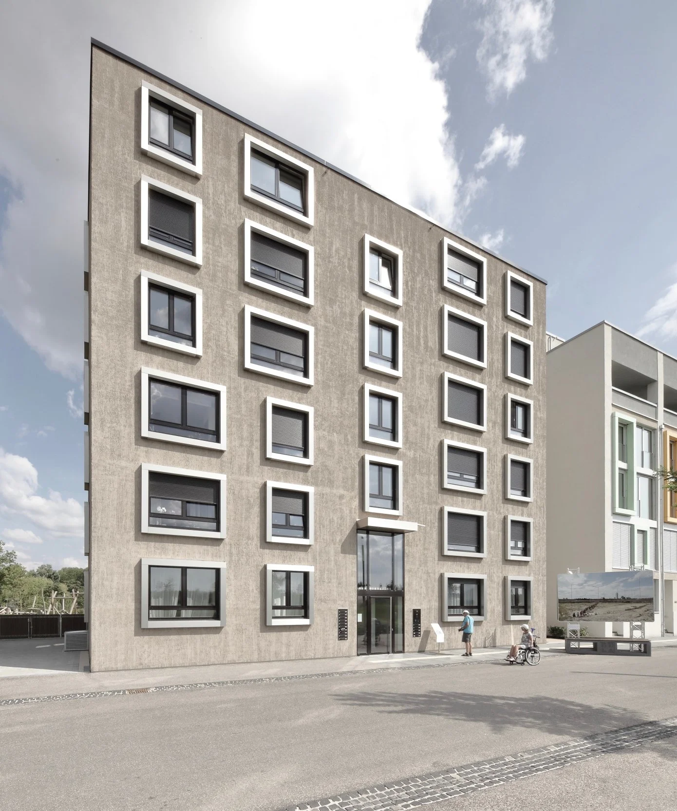 wohnhaus BUGA - Modernes mehrstöckiges Wohnhaus mit weißen Fensterrahmen und rollbaren Fensterläden, Eingangsbereich mit Glasfront, Leute im Vorfeld, Radfahrer vor dem Haus, ein Computerbildschirm zeigt eine Landschaftsbzeichnung.