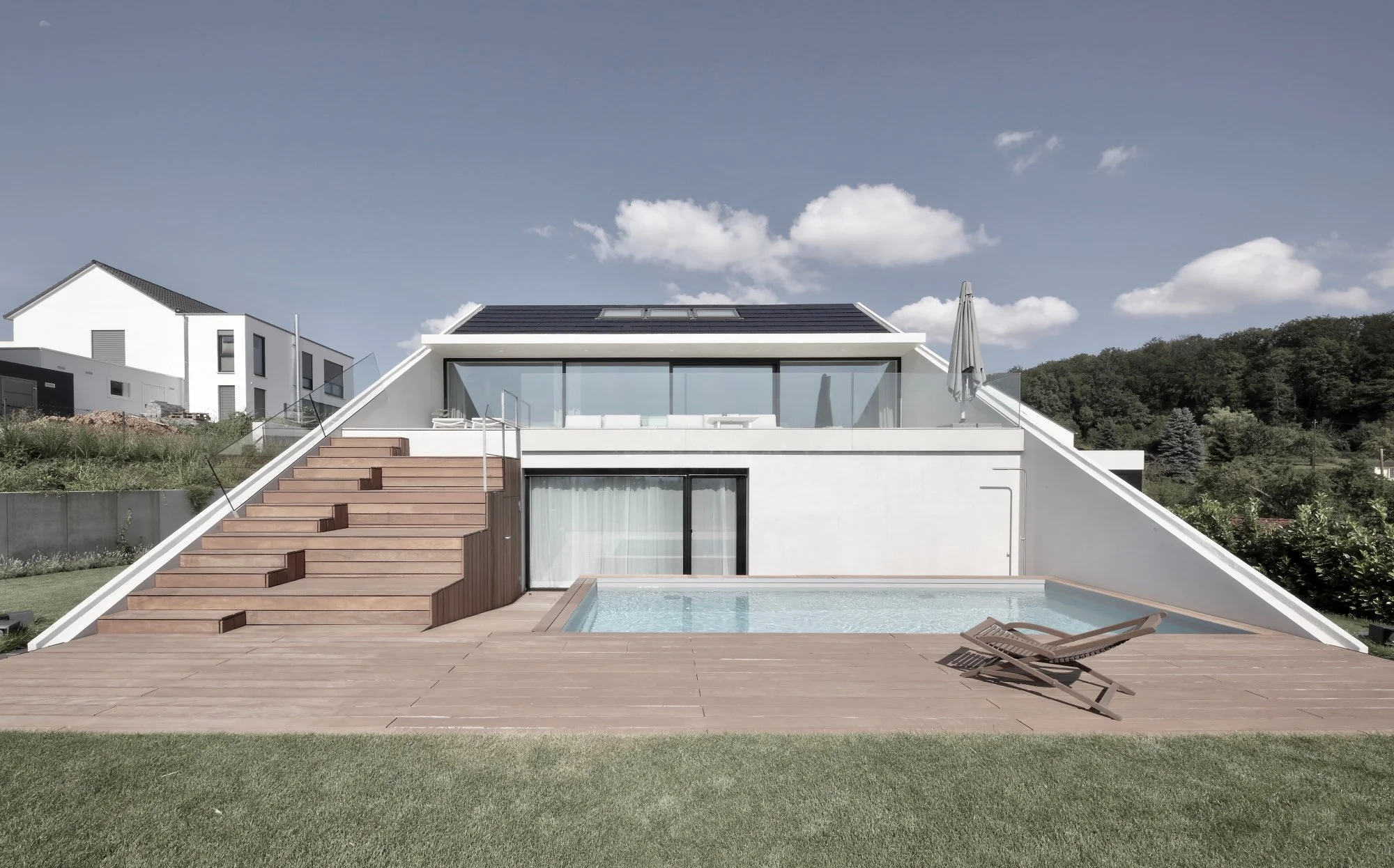 haus CC - Modernes weißes Haus mit Dachfenstern, Terrasse, Pool und Liegen, umgeben von grünem Garten und blauen Himmel.