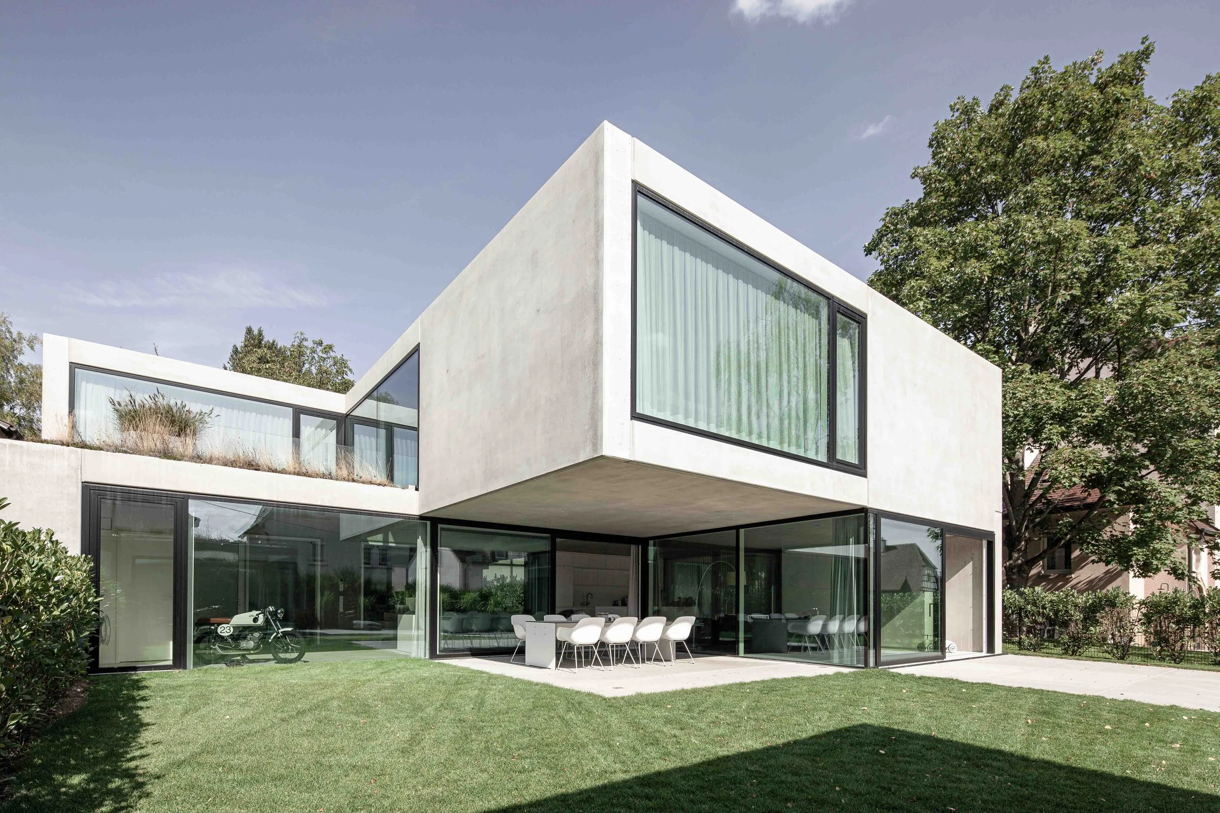 haus vO - Modernes, kubisches Haus mit großen Fensterflächen, umgeben von einem gepflegten Garten und einem Baum auf der rechten Seite, bei sonnigem Himmel.