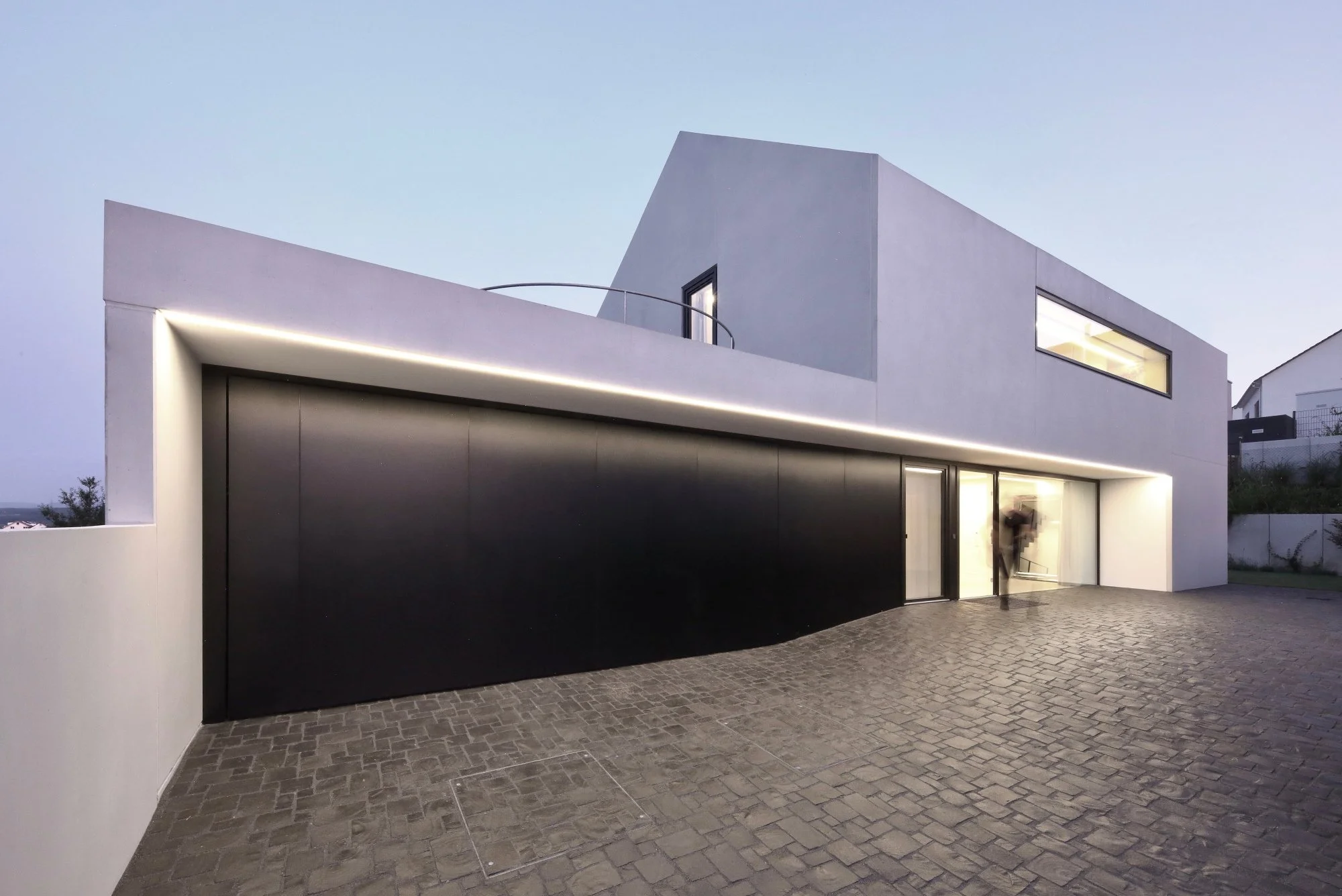 haus CC - Modernes zweistöckiges Haus mit weißen und schwarzen Wänden, große Fenster, Innenbeleuchtung sichtbar, gepflasterte Außenfläche bei Dämmerung.