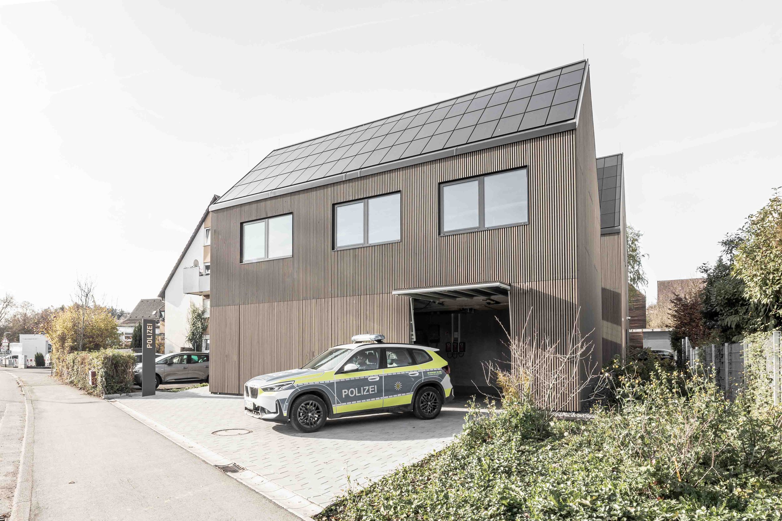 polizeiposten LE - Mehrstöckiges Gebäude mit Holzverkleidung und Solarpanelen auf dem Dach, Polizeifahrzeug vor dem Gebäude und einige Bäume und Pflanzen im Vordergrund.