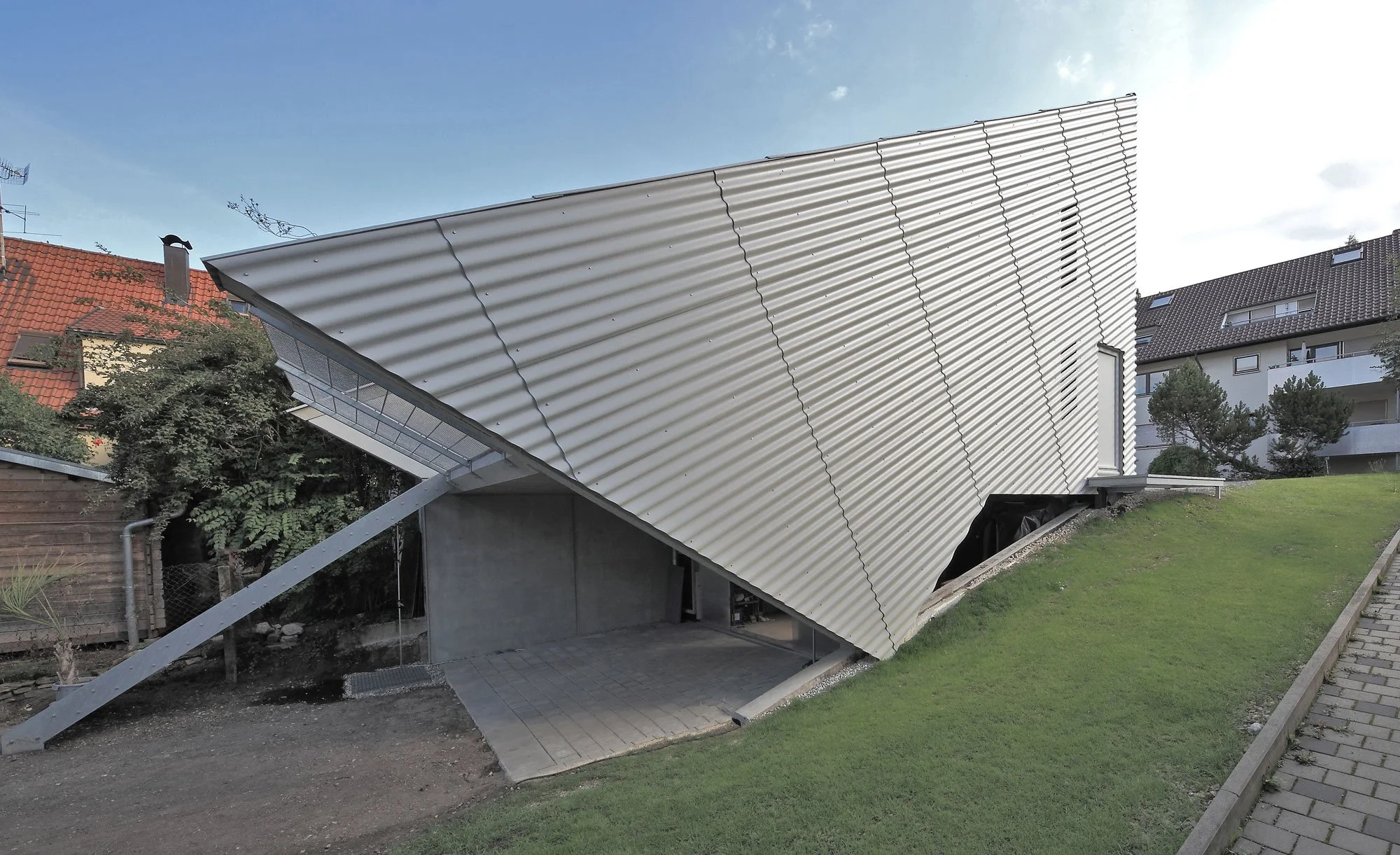 haus SO - Ein modernes Haus mit weit geneigtem, silbernem Metalldach, das die Architektur des Gebäudes betont, in einem Wohngebiet mit anderen Häusern im Hintergrund und einem gepflegten Rasen im Vordergrund.