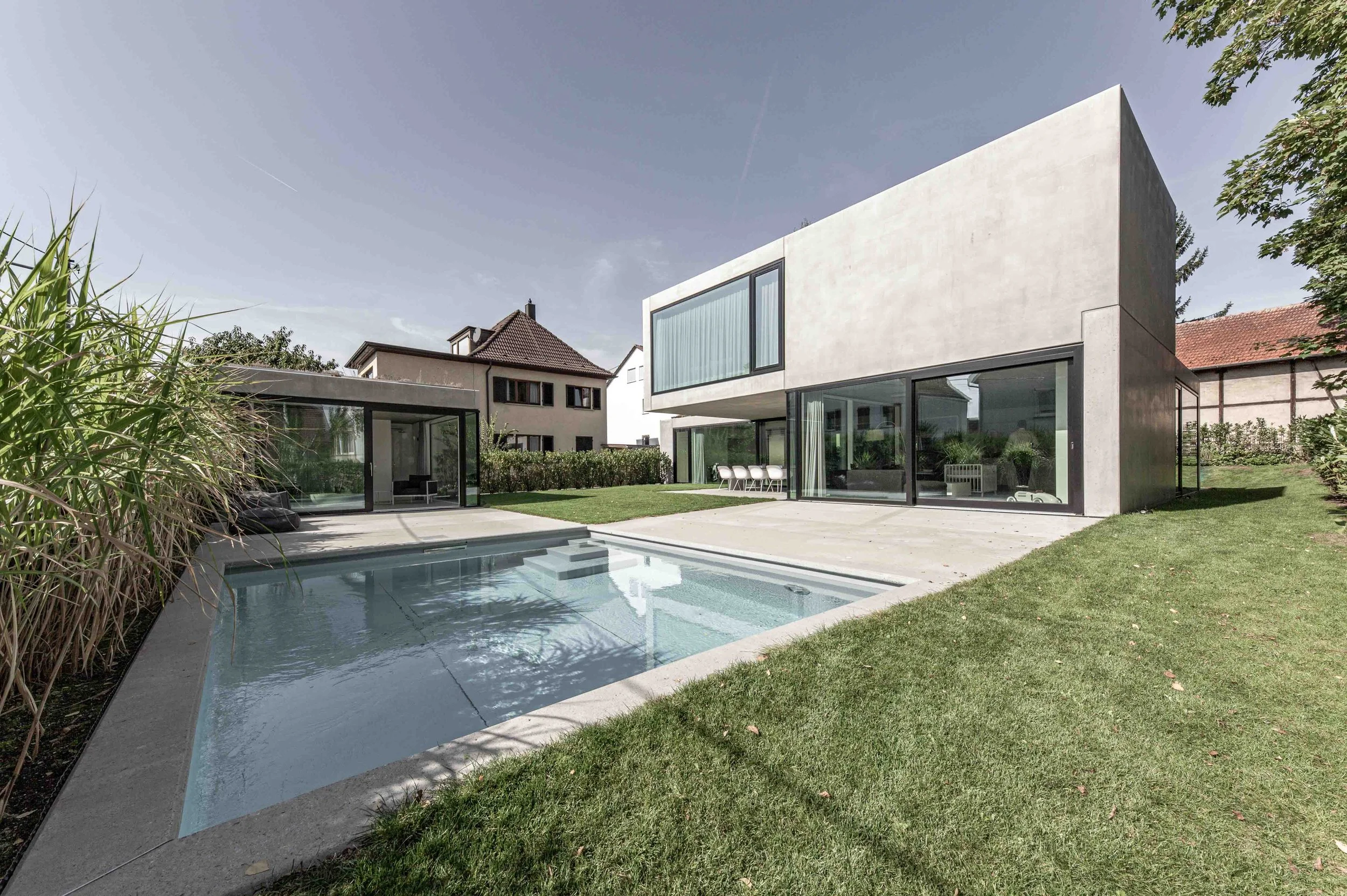 haus vO - Moderner zweigeschossiger Betonbau mit großen Glasflächen im Garten, umgeben von Rasen und Bäumen, mit Pool im Vordergrund.