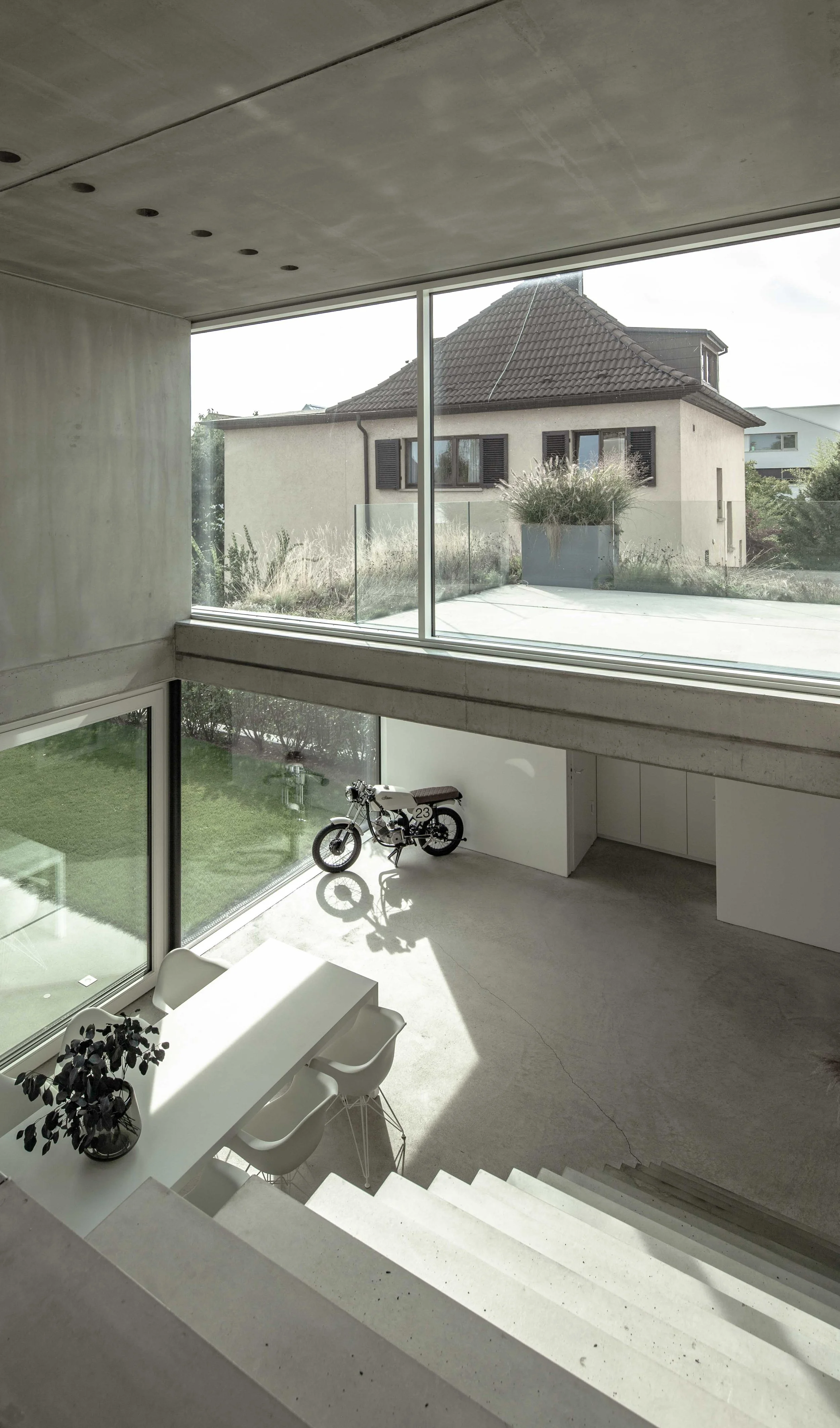 haus vO - Moderne Inneneinrichtung mit großen Fenstern, einem weißen Esstisch, weißen Stühlen, einer Pflanze im Topf, einem Rennrad, einer Treppe und einer Terrasse mit Blick auf ein Haus mit Dach und Schärfefenster.