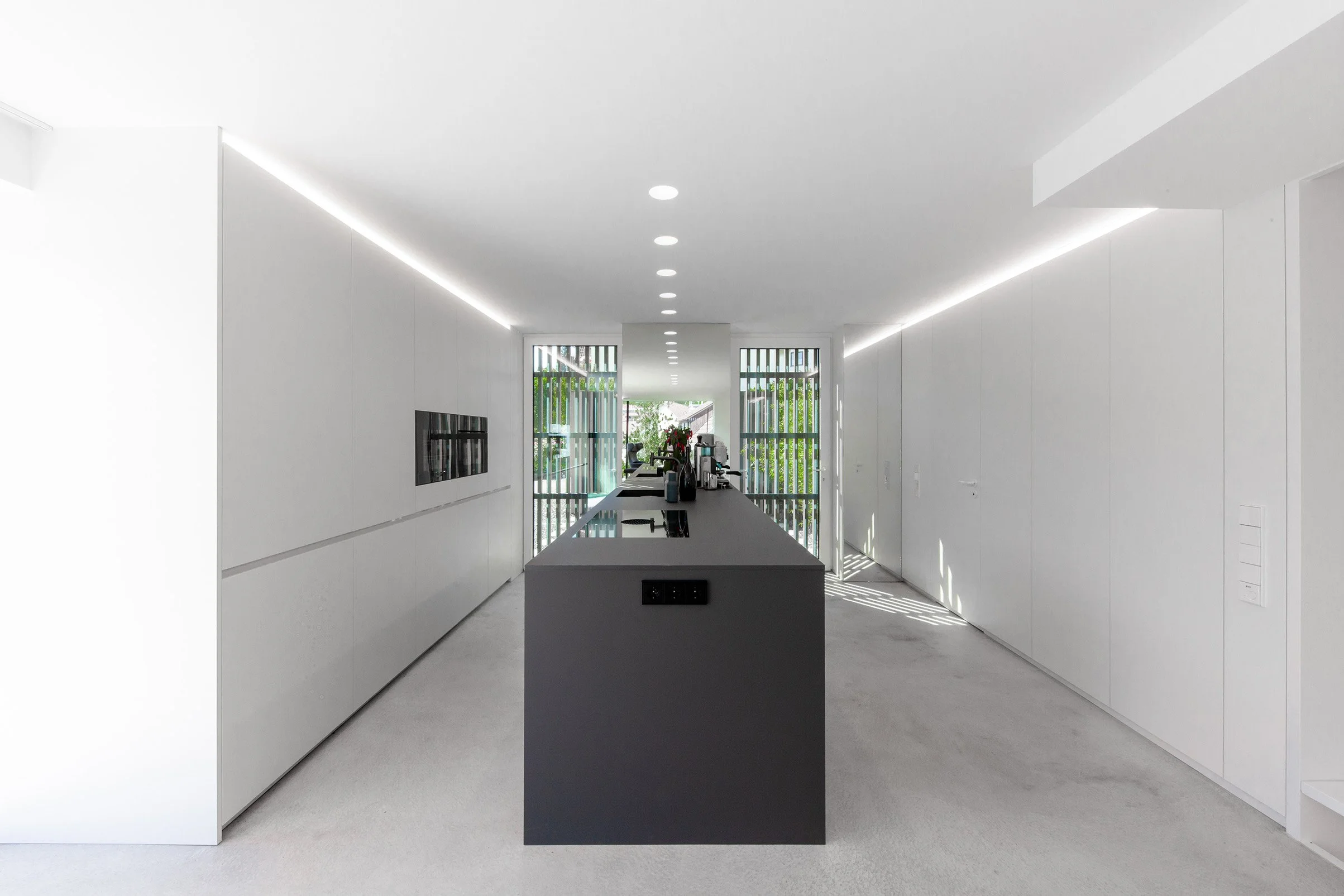 haus RU - Modern, minimalist Küche mit weißem Design, schwarzen Arbeitsflächen, großen Fenstern mit Gitterstäben, im Hintergrund sitzen Personen.