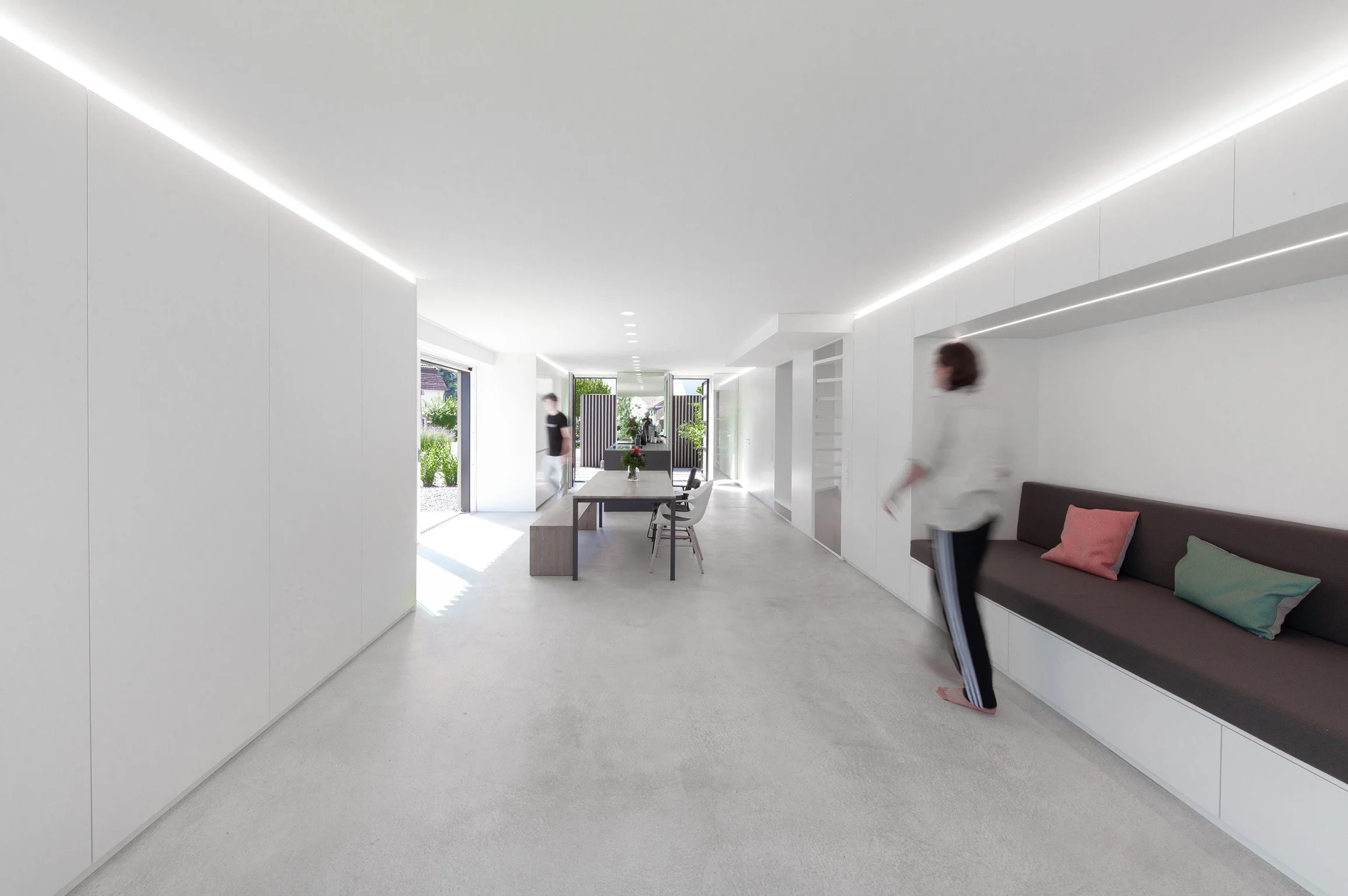 haus RU - Modernes, helles Wohnzimmer mit minimalistischer Einrichtung, grauem Sofa, bunten Kissen, großen Fenstern und offener Küche im Hintergrund.