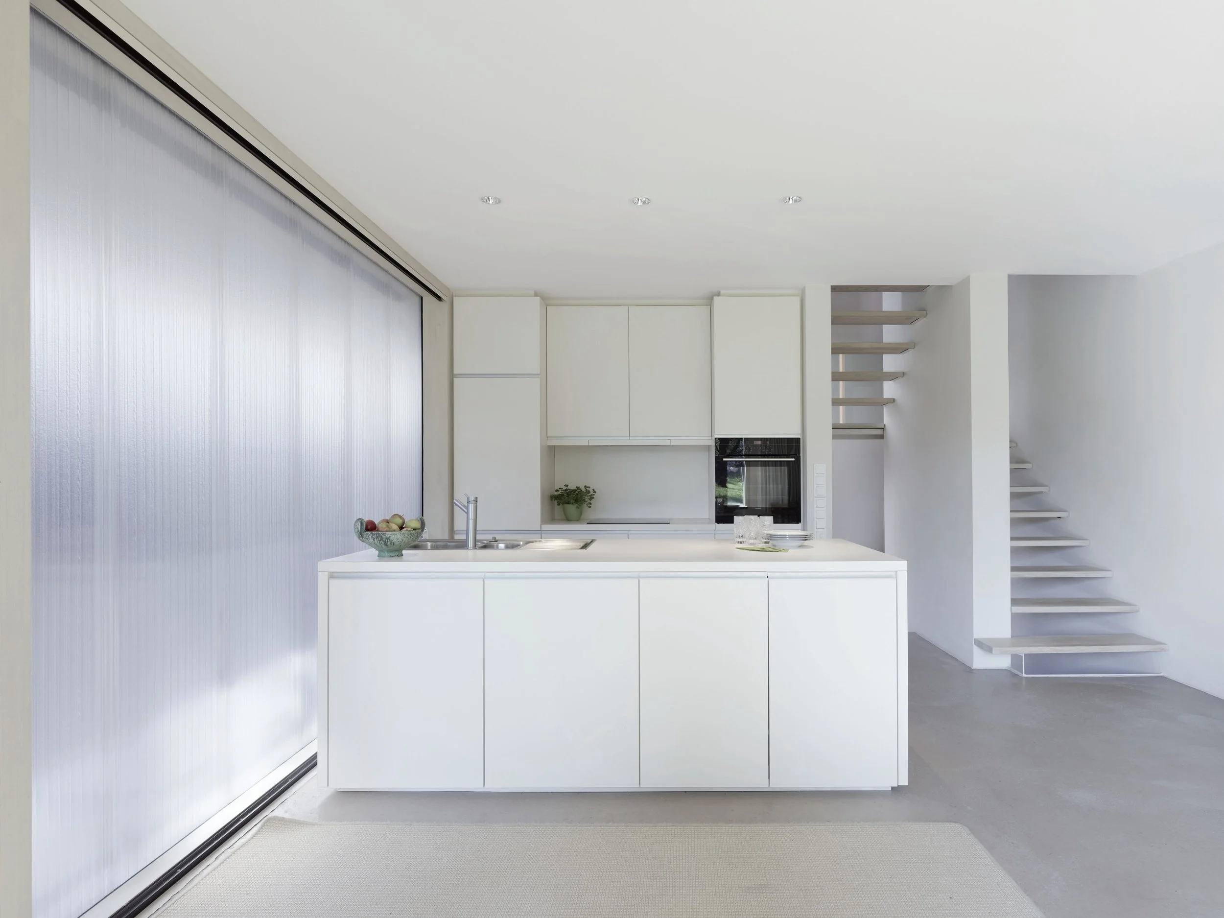 haus - B_Moderne weiße Küche mit Kücheninsel, Spüle, Einbauofen, Obstschale und angrenzender offener Treppe