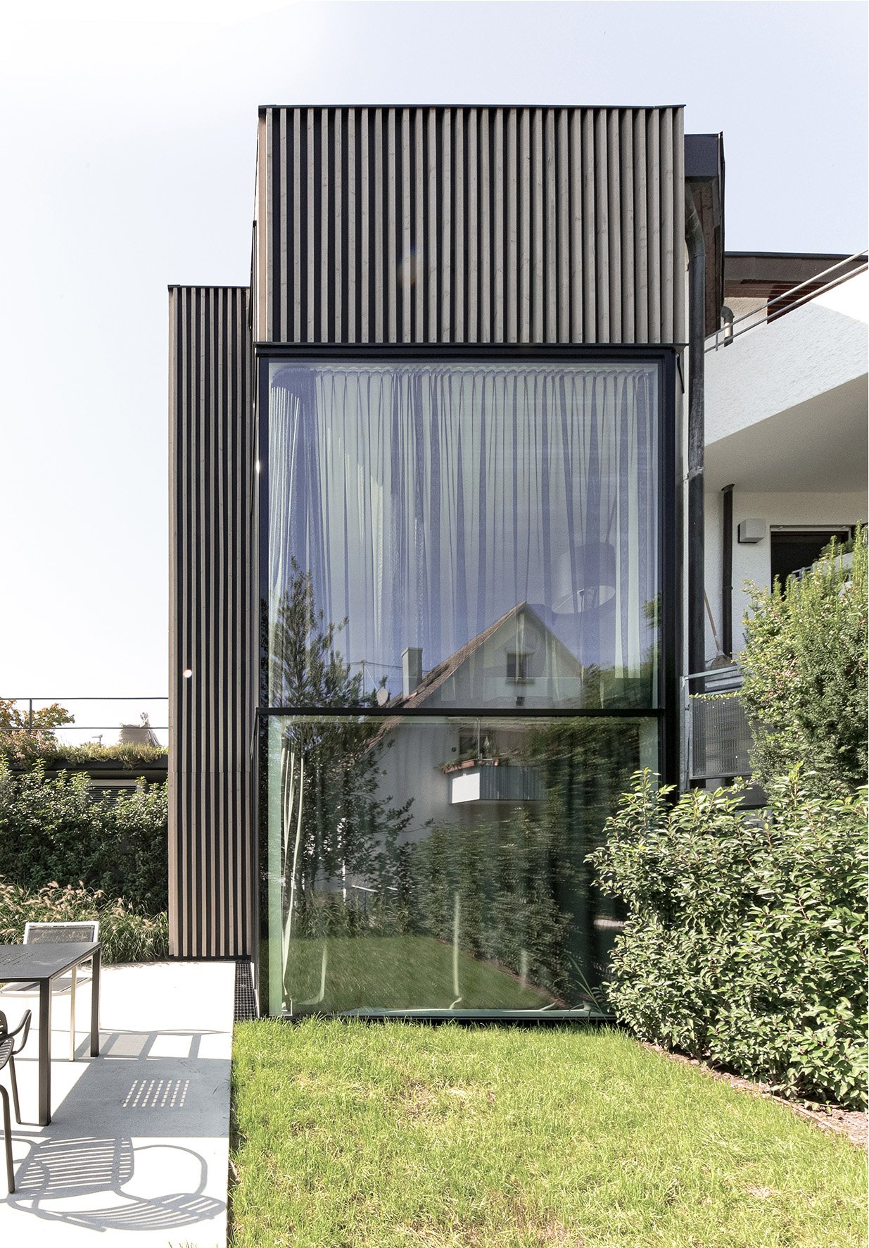 haus RU - Modernes Haus mit großen Fensterflächen und vertikalen Holzlamellen an der Fassade, Garten mit Sitzmöbeln und grüner Rasen im Vordergrund.
