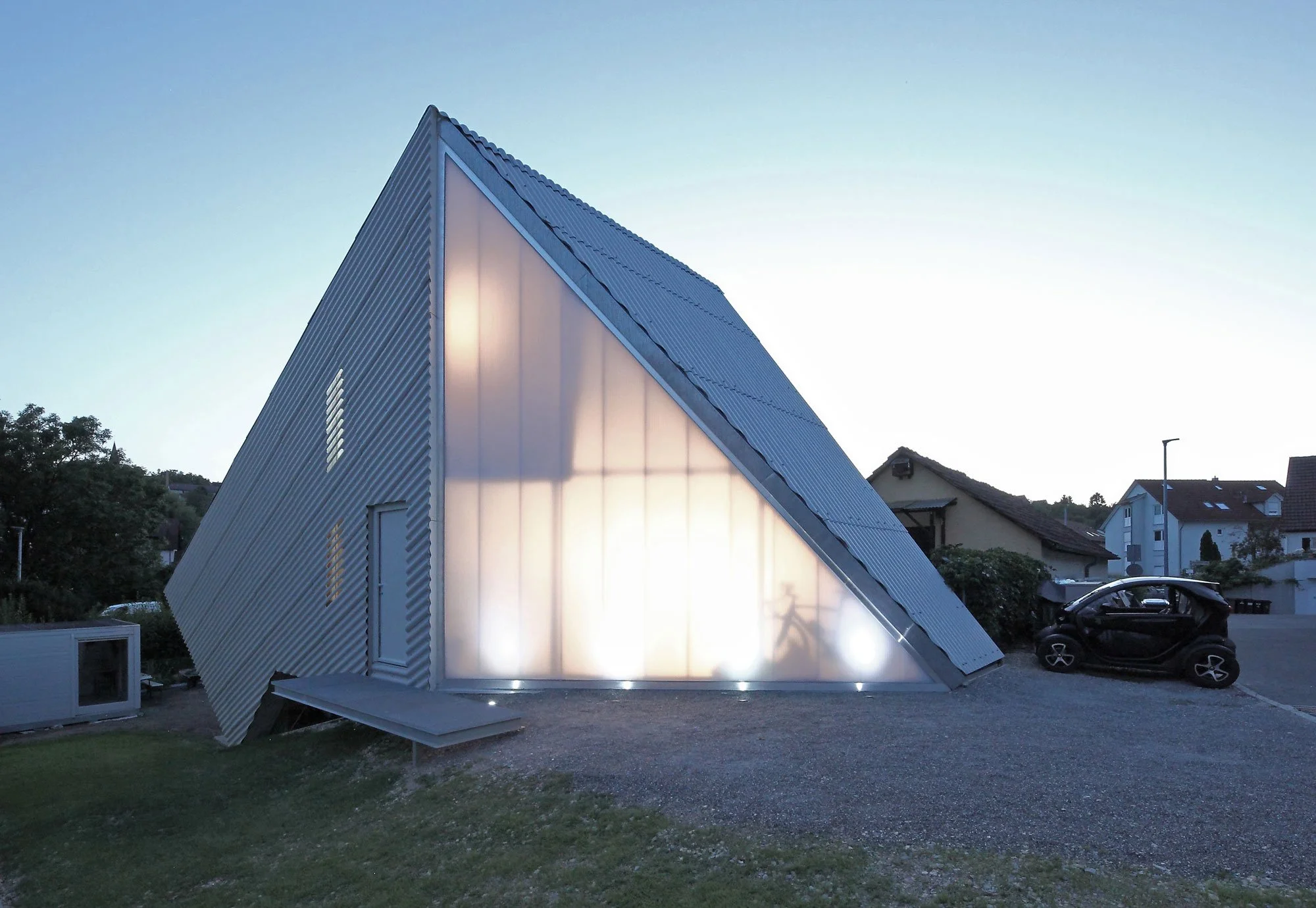 haus SO - Moderne, schiefe Gebäudearchitektur mit Metallfassade, das Gebäude ist nach oben geneigt und scheint schief zu stehen, Sonnenlicht scheint auf die Fassade, umgeben von Wohnhäusern und einem Auto in einer ruhigen Nachbarschaft.