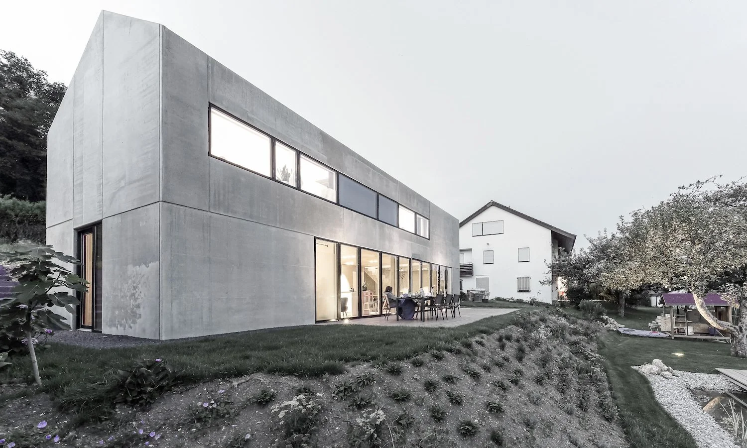 haus WÄ - Modernes zweistöckiges Haus mit großen Fenstern, umgeben von Garten mit Bäumen und Wiese, Abendstimmung