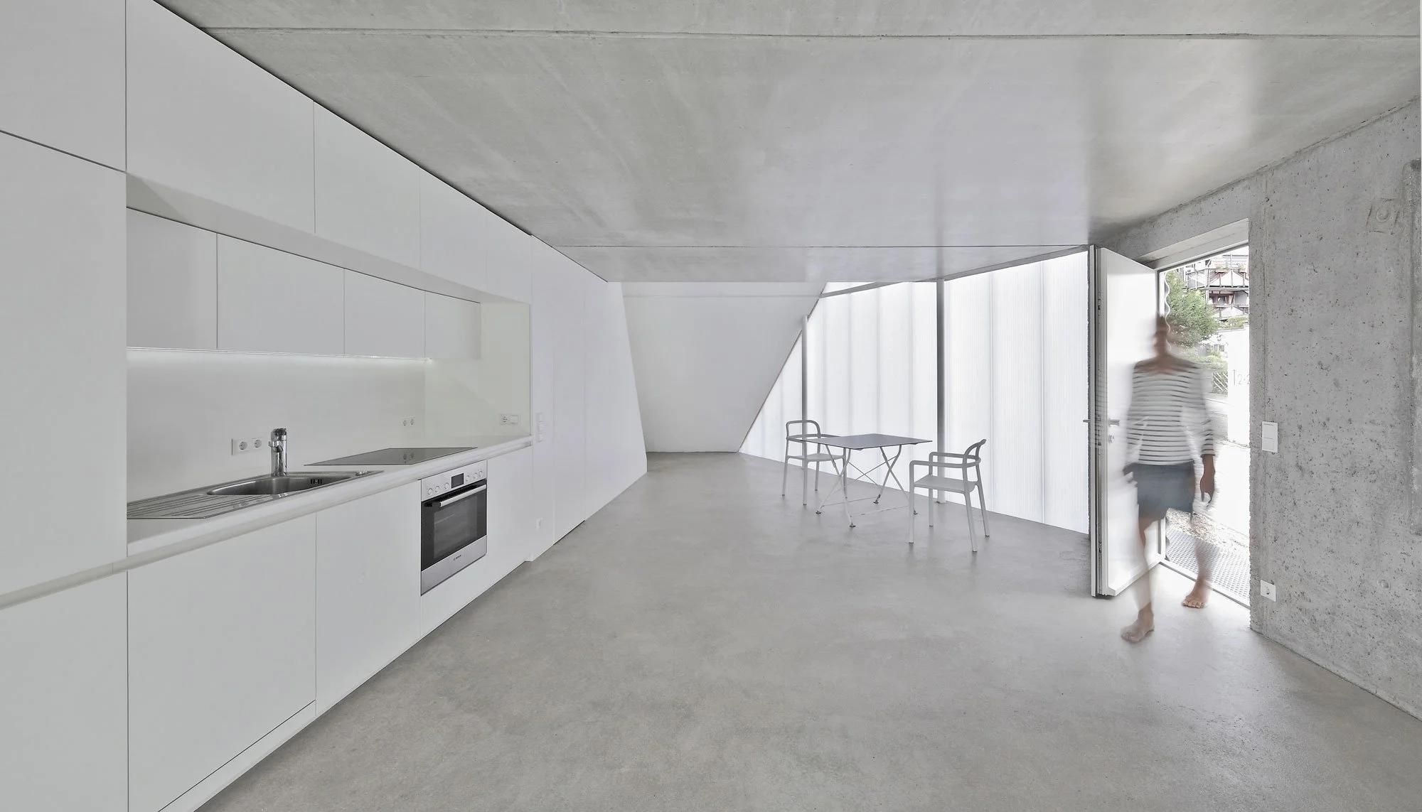 haus SO - Ein modern eingerichteter Raum mit weißen Wänden, einer Küchenzeile und einem Esstisch mit Stühlen. Große Fenster lassen natürliches Licht herein, und eine Person betritt den Raum.