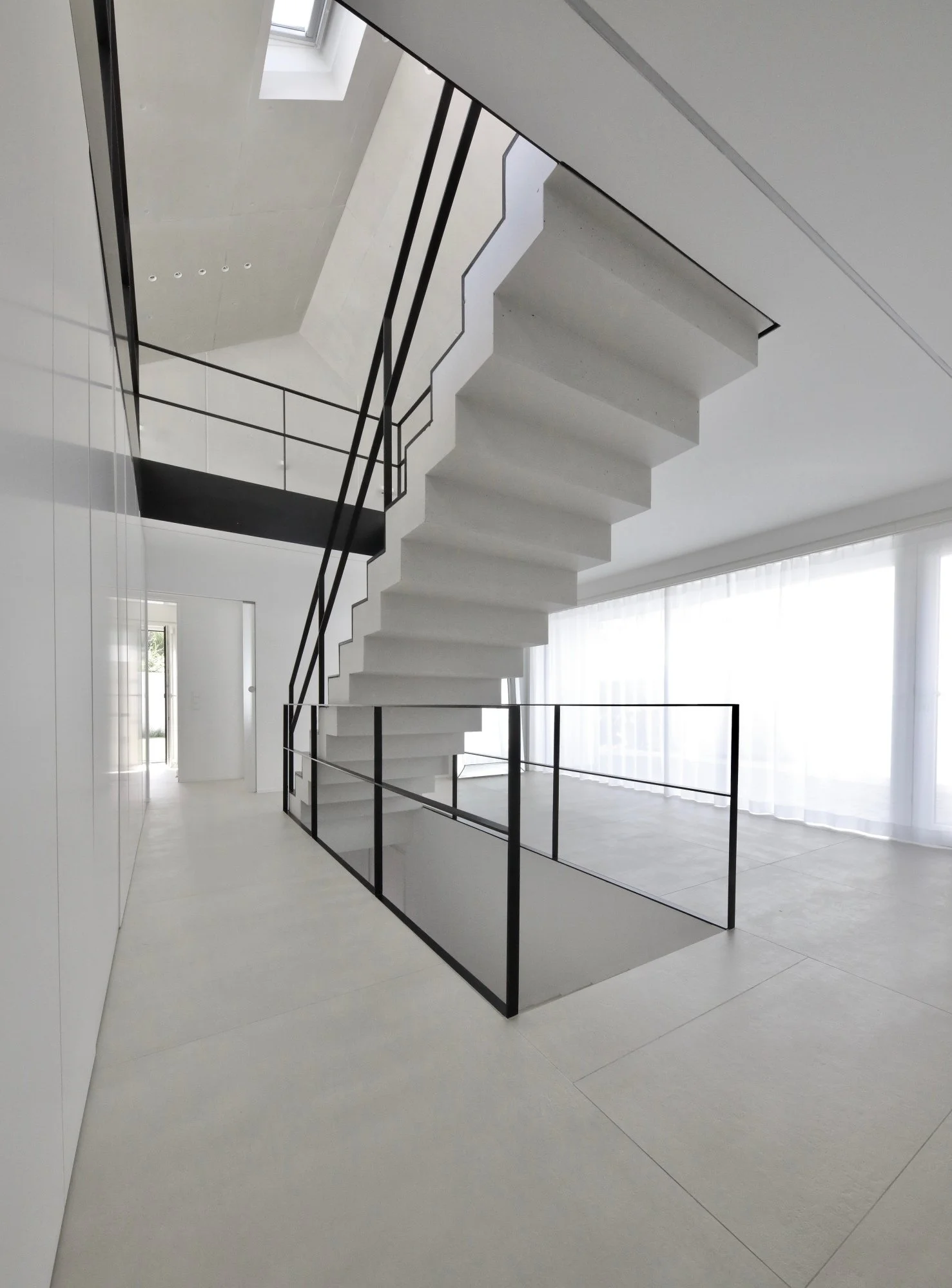 haus CC - Innenaufnahme eines modernen, minimalistisch gestalteten Treppenhauses mit weißen Wänden, schwarzem Geländer und großen Fenstern, die viel Tageslicht hereinlassen.