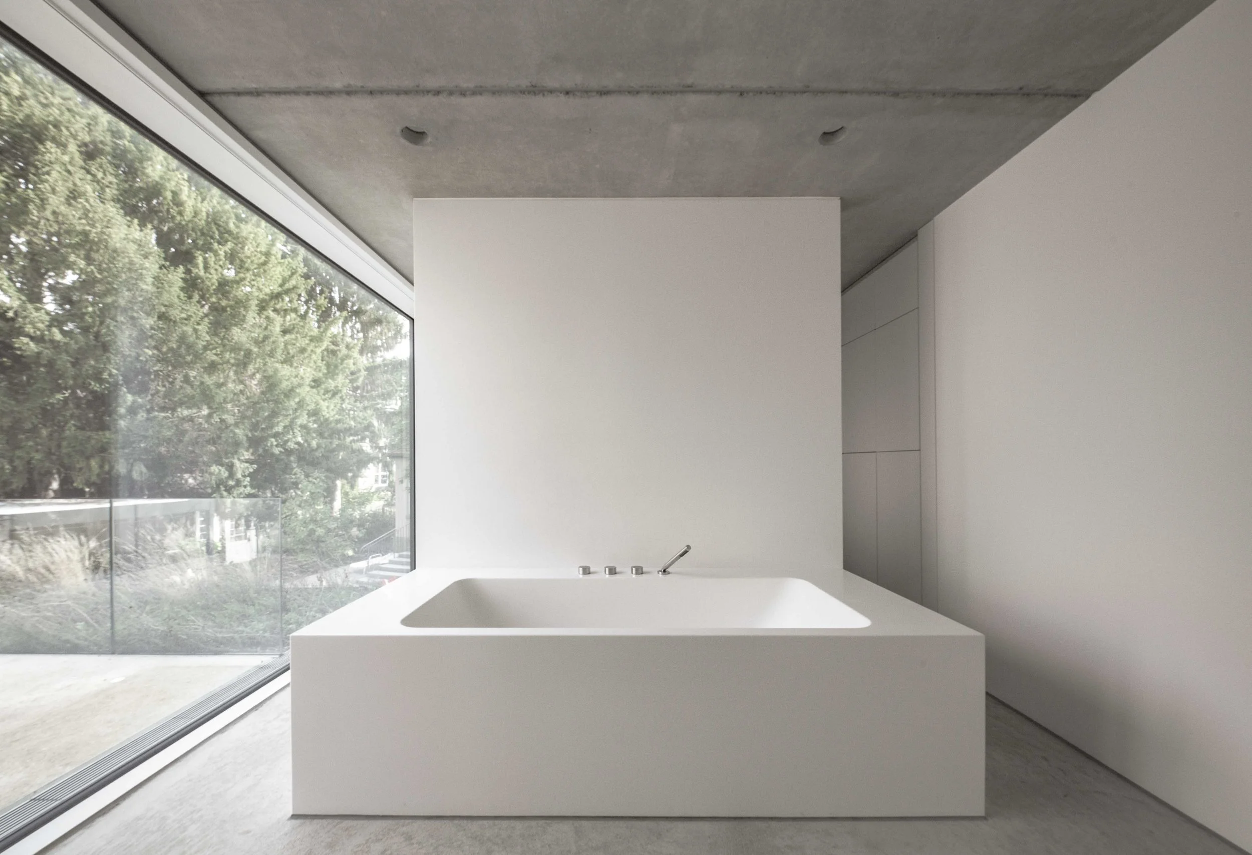 haus vO - Ein moderner, weißer Badezimmerbereich mit einer Eckbadewanne, große Fenster mit Blick auf Bäume im Freien, graue Decke und weiße Wände.