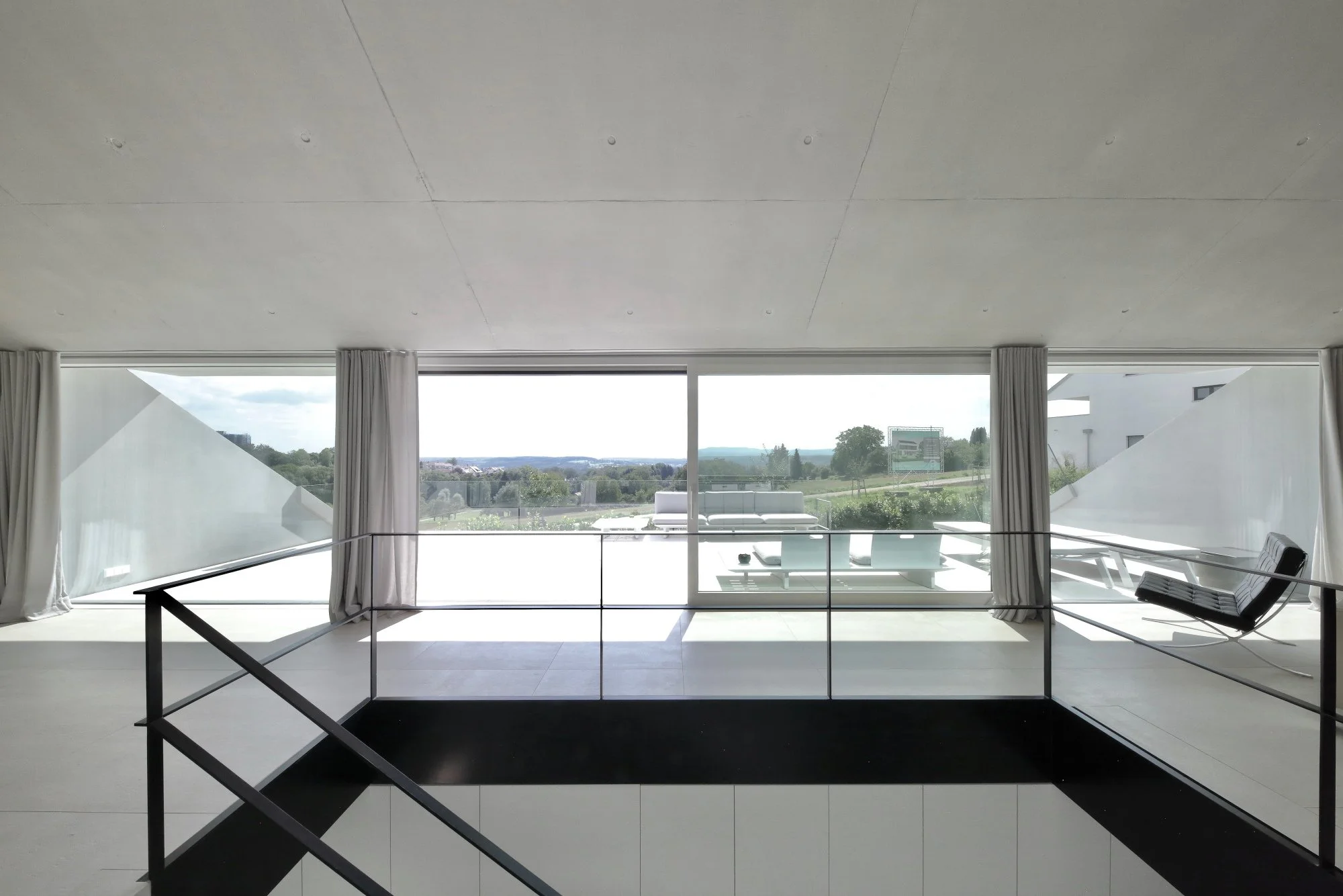 haus CC - Moderner Wohnraum mit großen Fenstern, Blick auf grüne Landschaft, schwarze Treppe im Vordergrund, weiße Möbel im Freien auf dem Balkon, klare Linien, helle Atmosphäre.