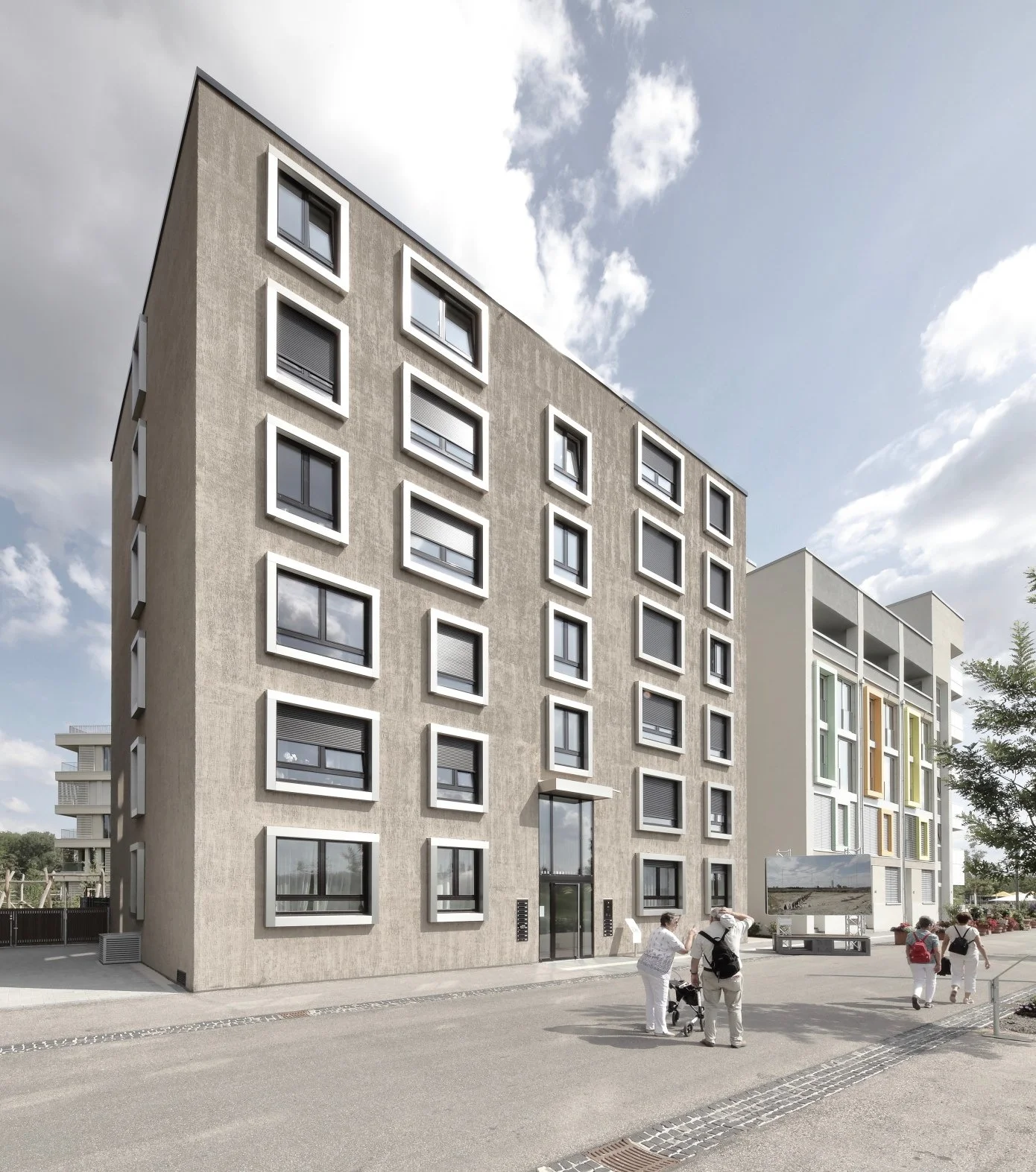 wohnhaus BUGA - Modernes mehrstöckiges Wohngebäude mit viereckigen Fenstern und Betonfassade, einige Menschen stehen vor dem Gebäude, im Hintergrund sind weitere Gebäude sichtbar, der Himmel ist teils bewölkt.