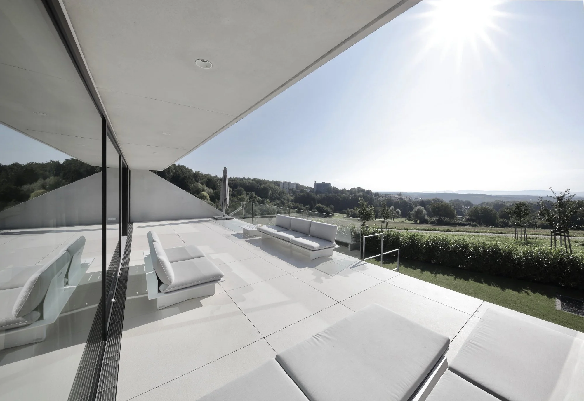 haus CC - Moderne Terrasse mit weißen Sitzmöbeln unter einem großen Balkon, mit Blick auf einen grünen Landschaft und einen klaren Himmel bei sonnigem Wetter.