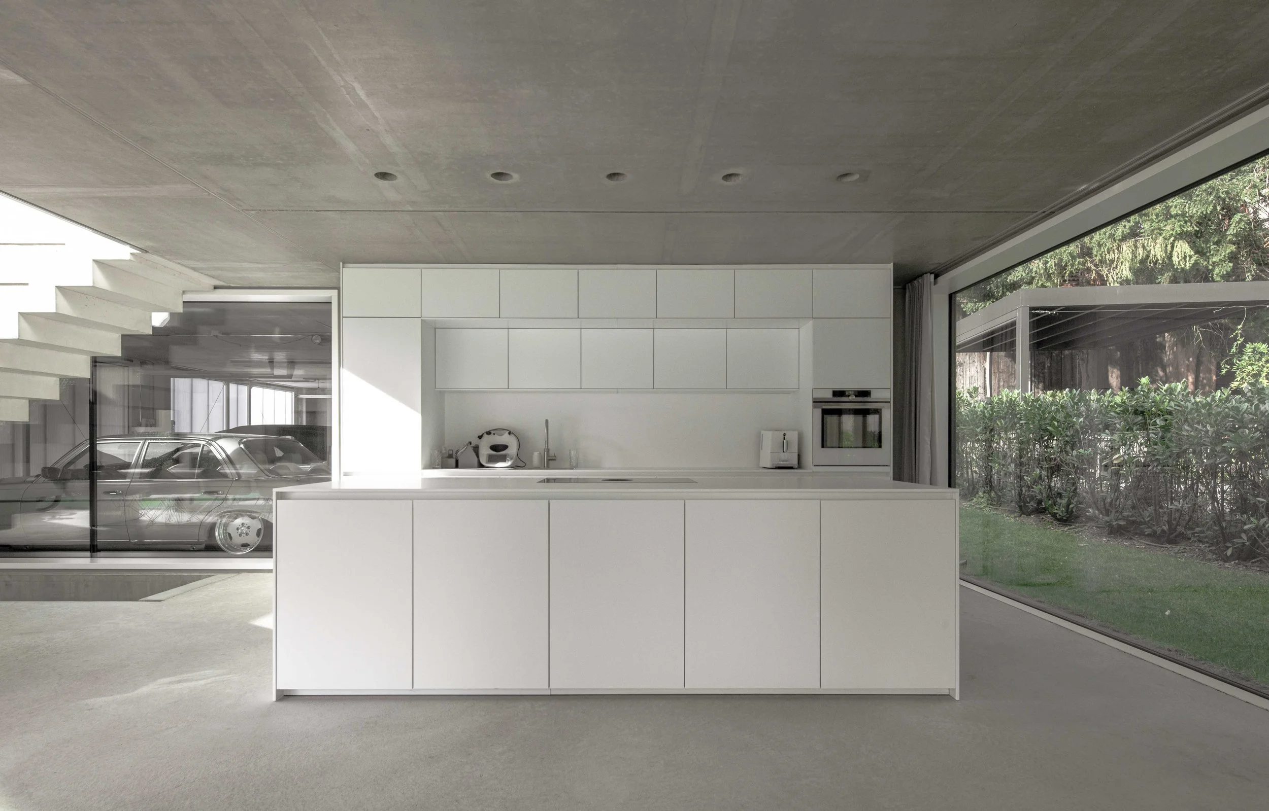haus vO - Moderne, minimalistische Küche mit weißen Schränken, eingebauten Elektrogeräten, großen Fenstern mit Ausblick in den Garten, Carport mit geparktem Auto, grauem Boden und Dach, viel natürlichem Licht.