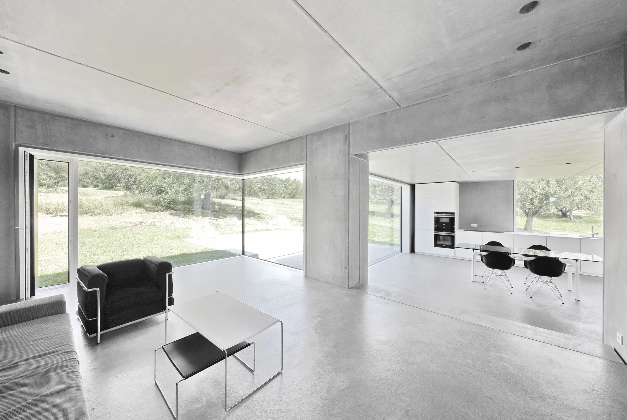 haus SG - Moderner, minimalistischer Wohnraum mit großen Fenstern, schwarzer Sitzecke, weißer Küche und offenem Grundriss.