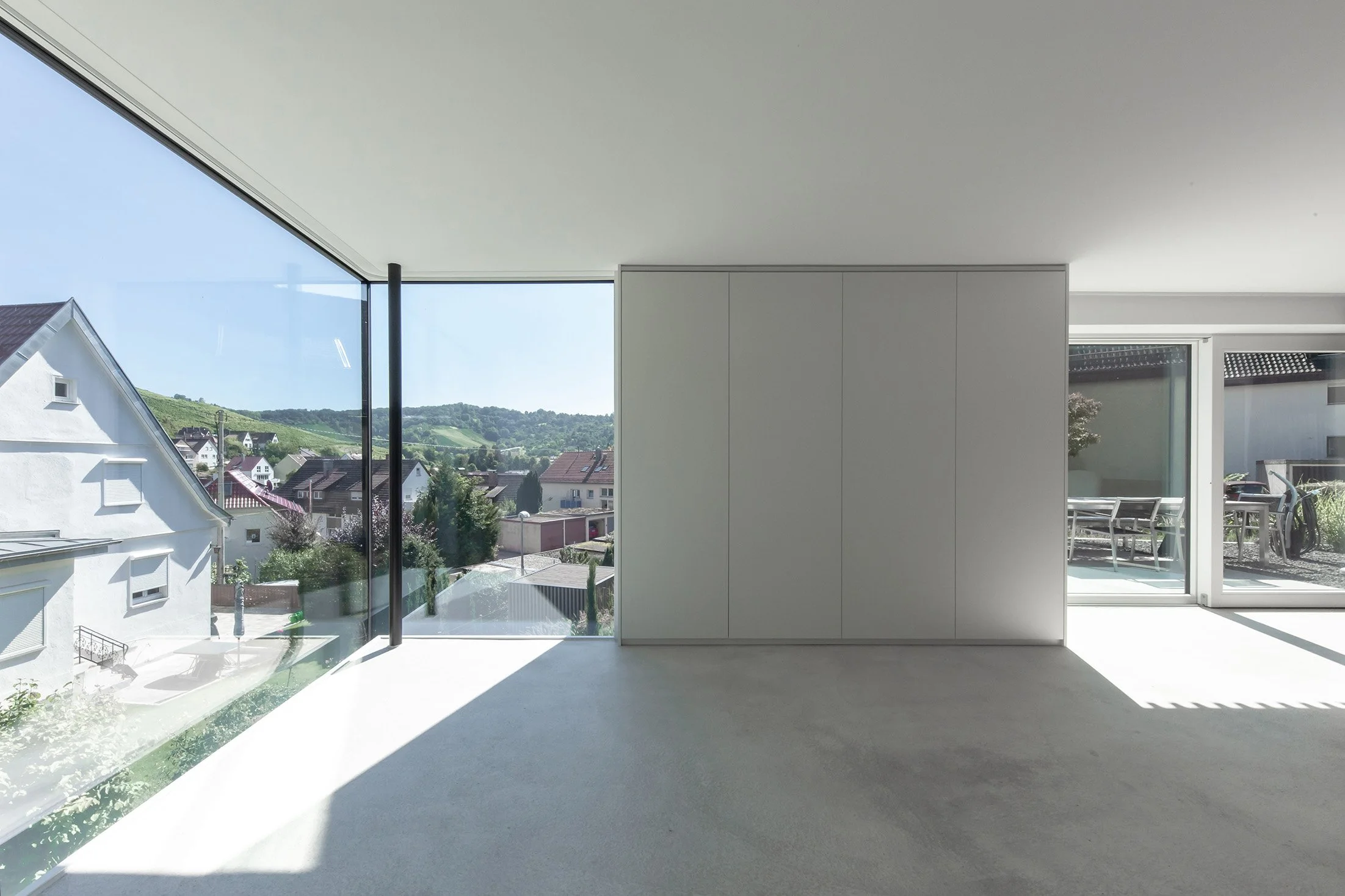 haus RU - Innenraum eines modernen Hauses mit großen Glasfenstern, Blick auf eine ländliche Gegend mit Häusern, Hügeln und Bäumen im Hintergrund, klarer Himmel, minimalistischer Innenraum mit weißen Wänden und unbedecktem Boden.