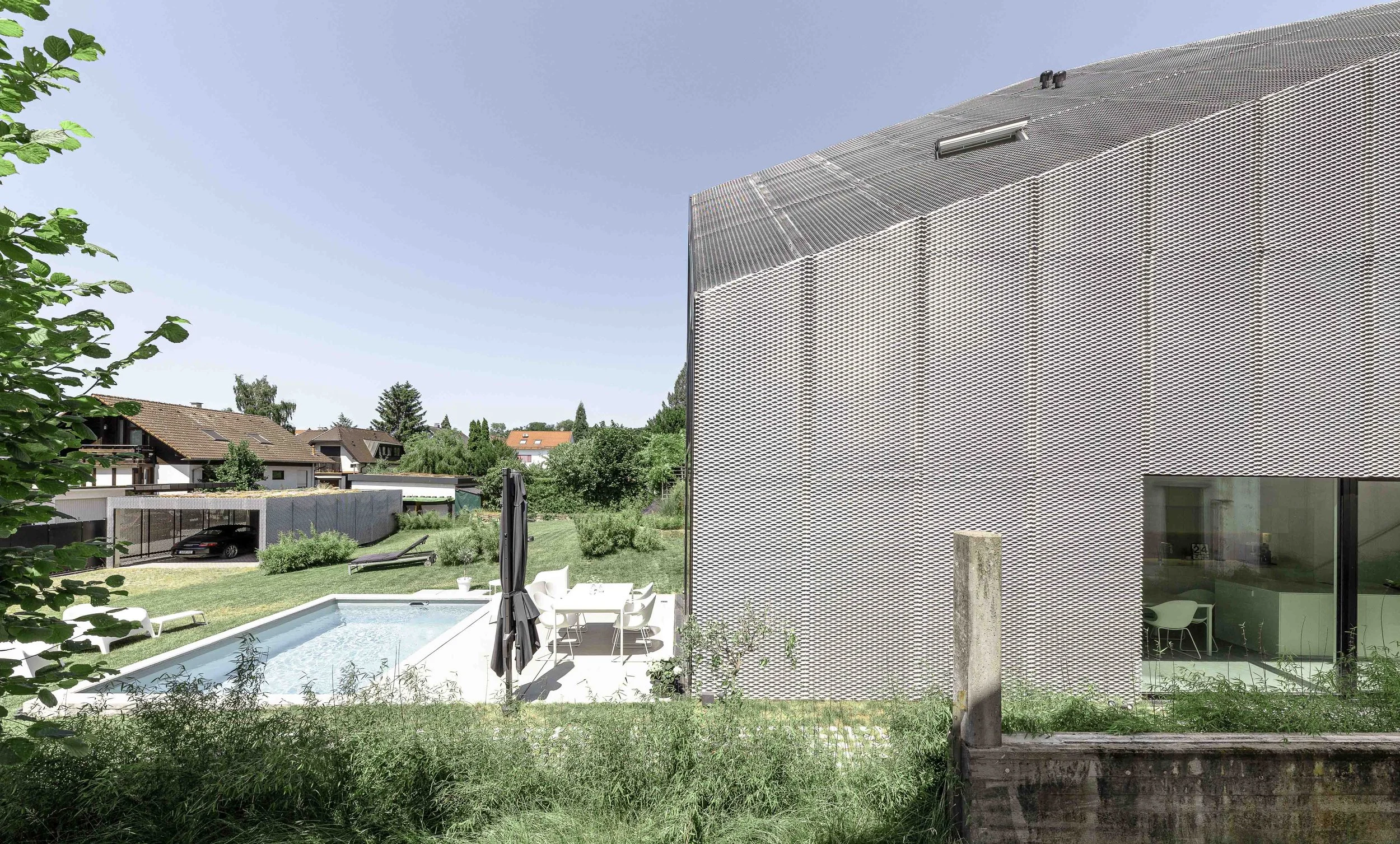 Holzterrasse mit Pool, umgeben von Garten und modernen Häusern, im Hintergrund blauer Himmel.