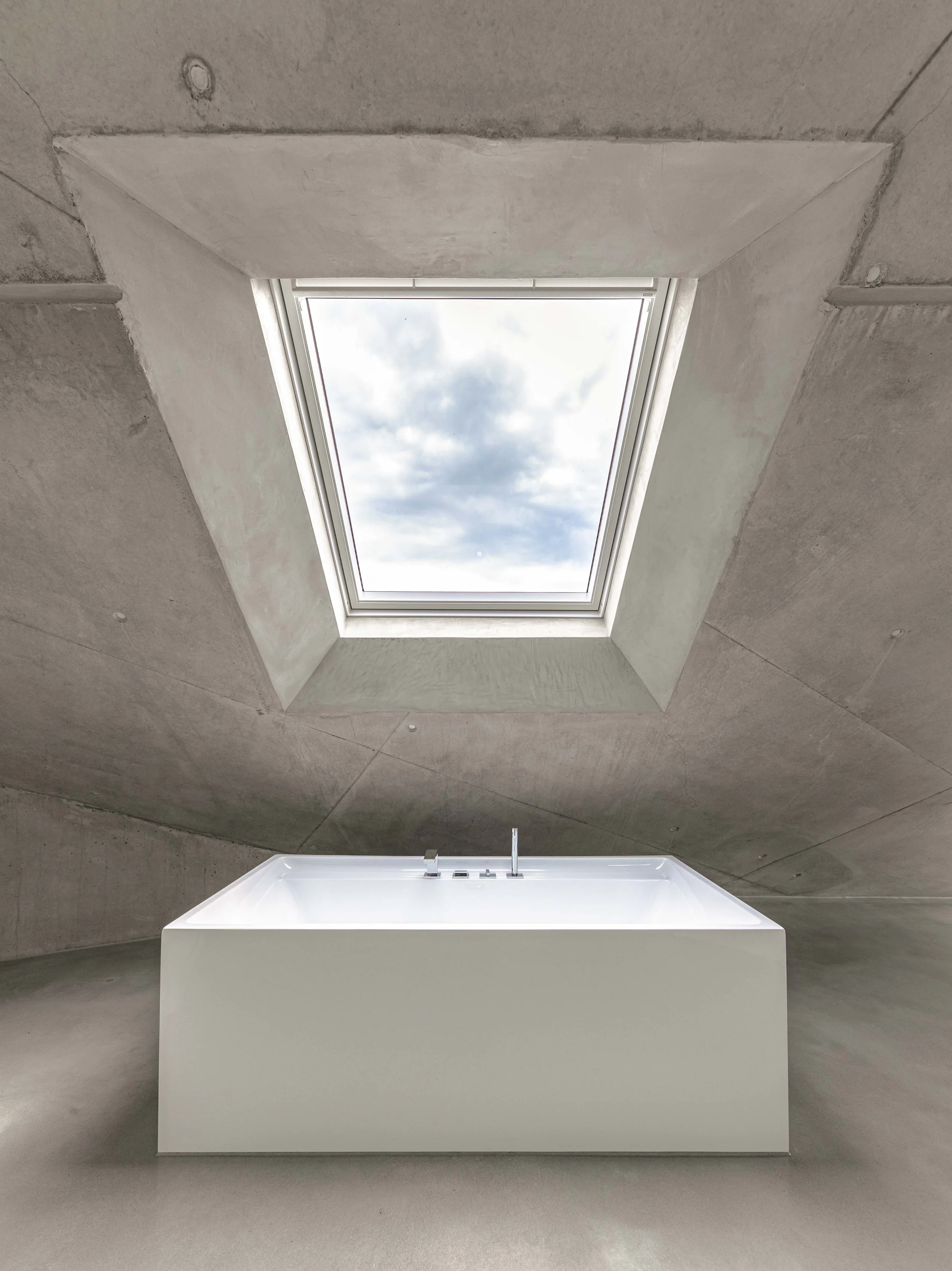 Innenansicht eines Raums mit einer großen Dachluke, die den Himmel mit Wolken sichtbar macht, und einer weißen Badewanne im Vordergrund auf glattem grauem Boden, die an einer modernen, minimalistischen Architektur beteiligt ist.