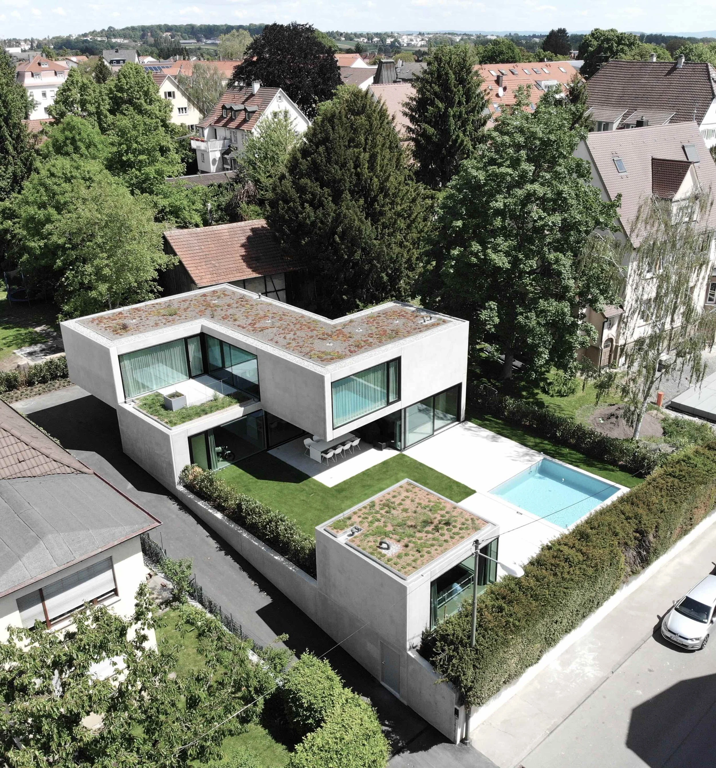 haus vO - Ein moderner, weißer Gebäudekomplex mit großen Fenstern, umgeben von Bäumen und Grünflächen, mit einem Pool im Garten, in einem Wohngebiet mit anderen Einfamilienhäusern im Hintergrund.