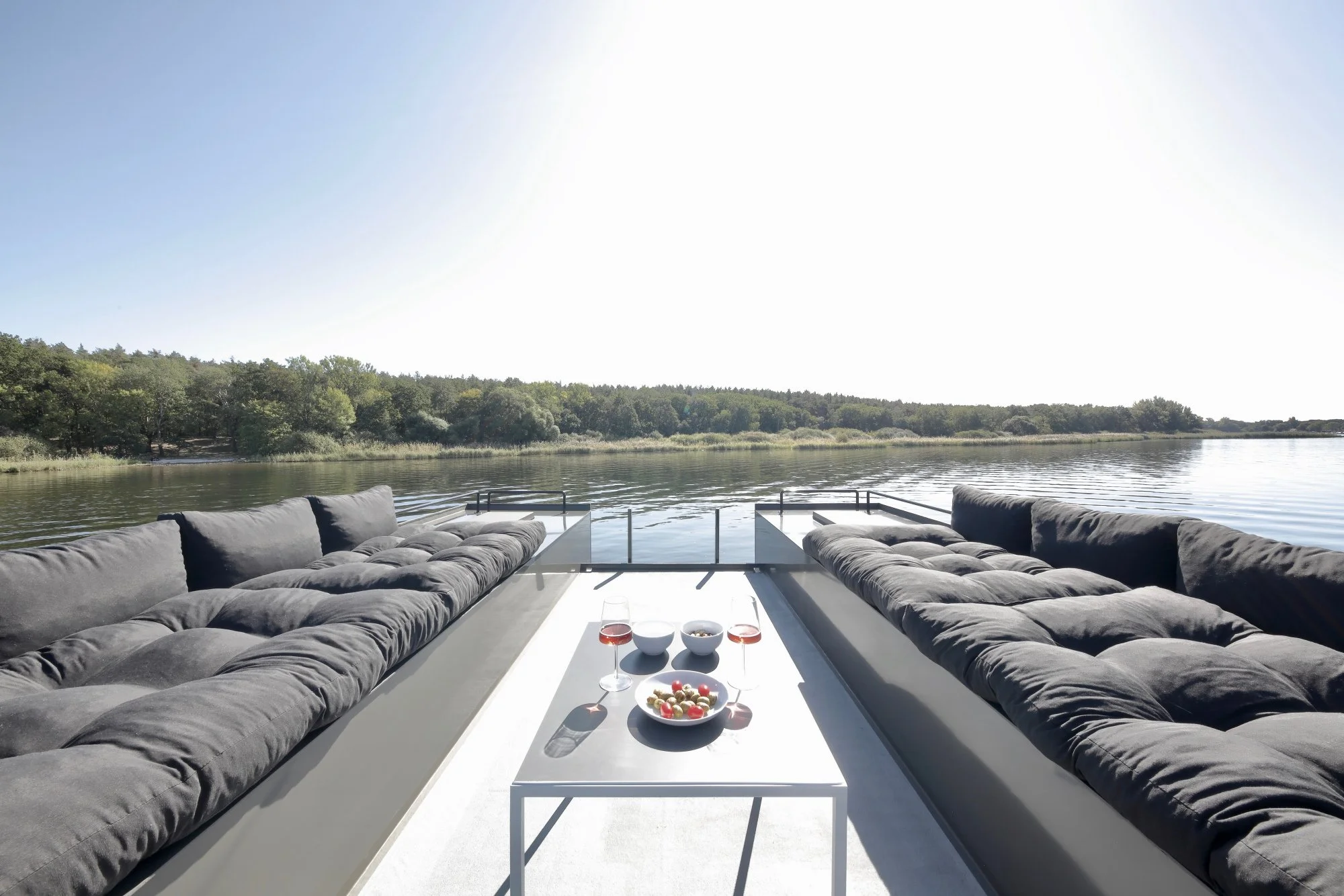 Außerhalb auf einem Boot oder Hausboot sitzt man auf schwarzen gepolsterten Sofas, vor einem weißen Tisch mit Getränken, Snacks und Chips, mit Blick auf einen Fluss und einen bewaldeten Horizont.