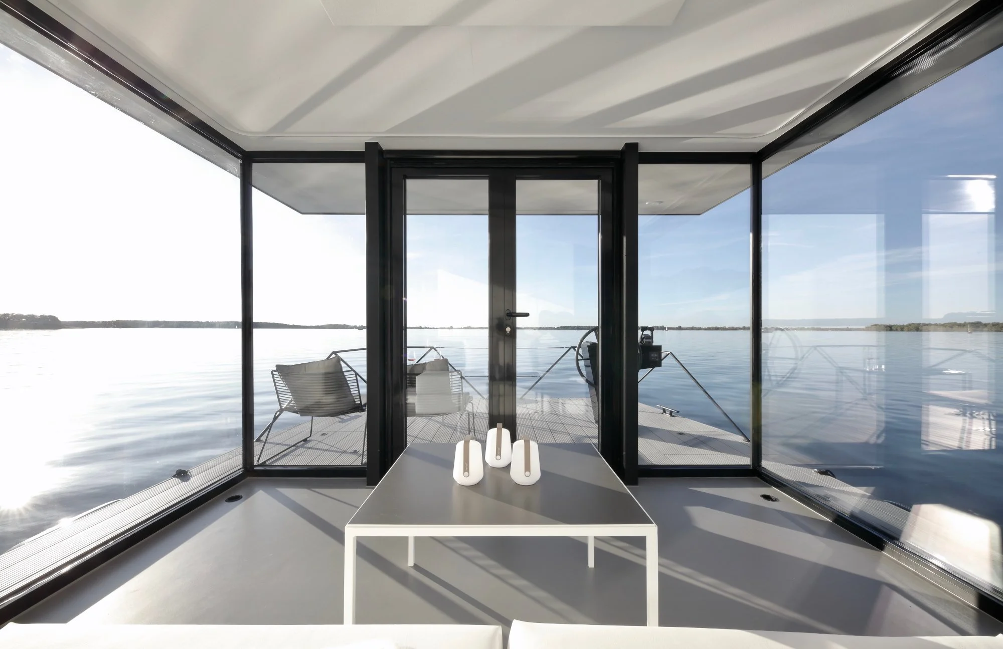 Innenansicht eines modernen Hauses mit großen Glasscheiben, Blick auf einen See, zwei Stühle und eine Tischlampe auf einer weißen Tafel