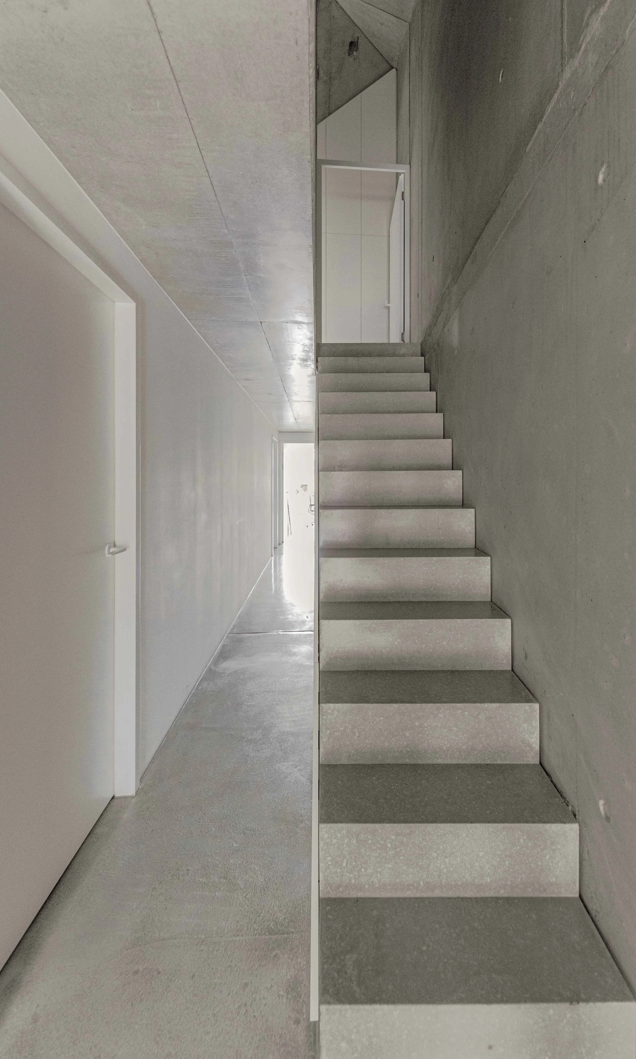 Innenansicht eines Gebäudes mit Treppe und weißem, modernen Design, grauer Wand und Betonboden.