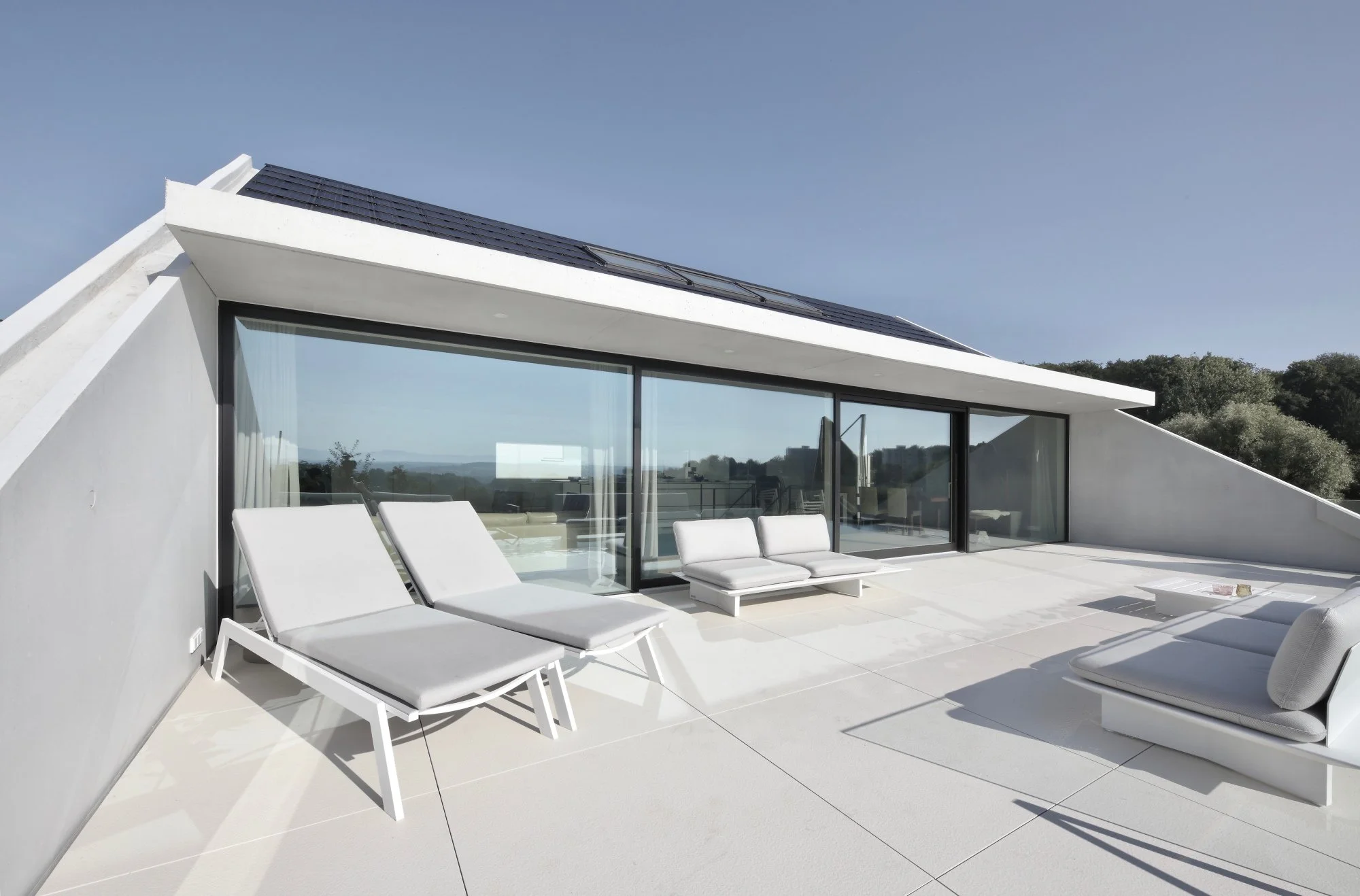 haus CC - Moderner weißer Balkon mit weißen Sonnenliegen, Sitzsäcken und großem Glasfenster, Blick auf eine ländliche Landschaft und blauen Himmel.