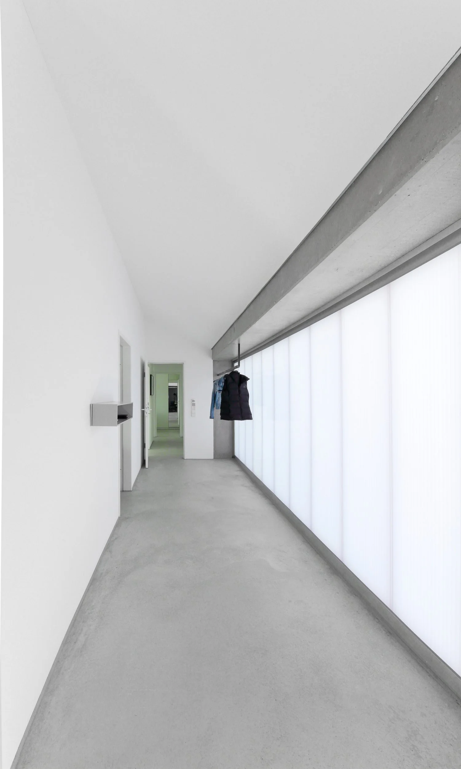 haus WÄ - Korridor in einem modernen Gebäude mit weißen Wänden, einem großen beleuchteten Fenster an der rechten Seite und Jacken, die an einer Stange hängen.