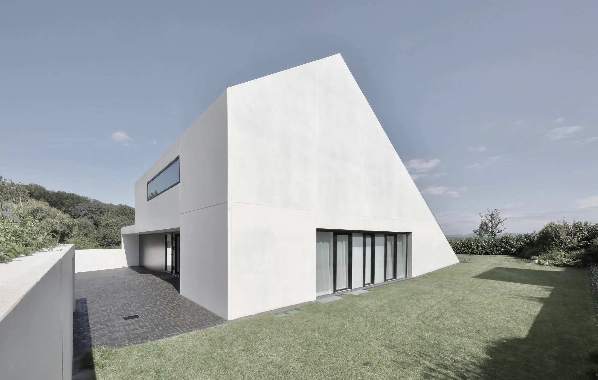 haus CC - Ein modernes weißes Haus mit großen Fenstern, umgeben von einem gepflegten Garten und Bäumen, bei blauem Himmel.