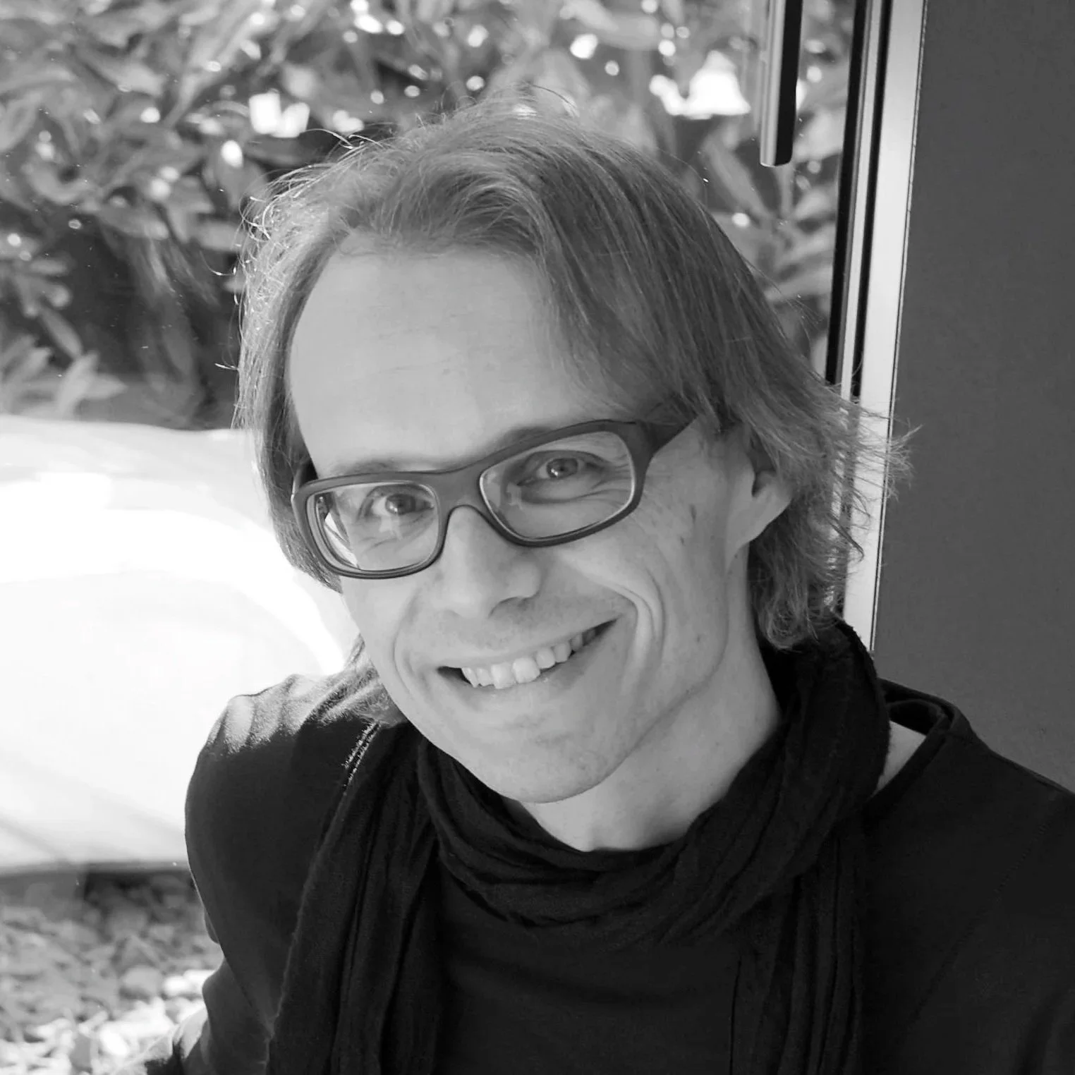 Ein Mann mit Brille, lächelnd, kurze Haare, trägt eine schwarze Jacke, im Hintergrund Pflanzen, schwarz-weiß Fotografie.