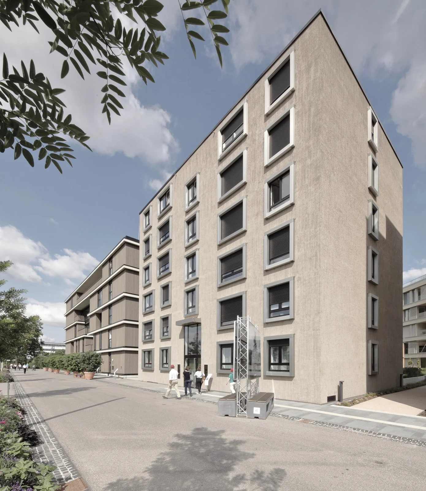 wohnhaus BUGA - Mehrstöckiges modernes Wohngebäude mit großen Fenstern und angelegtem Gehweg vor dem Gebäude, einige Fußgänger und Bäume im Vordergrund, blauer Himmel mit Wolken im Hintergrund.